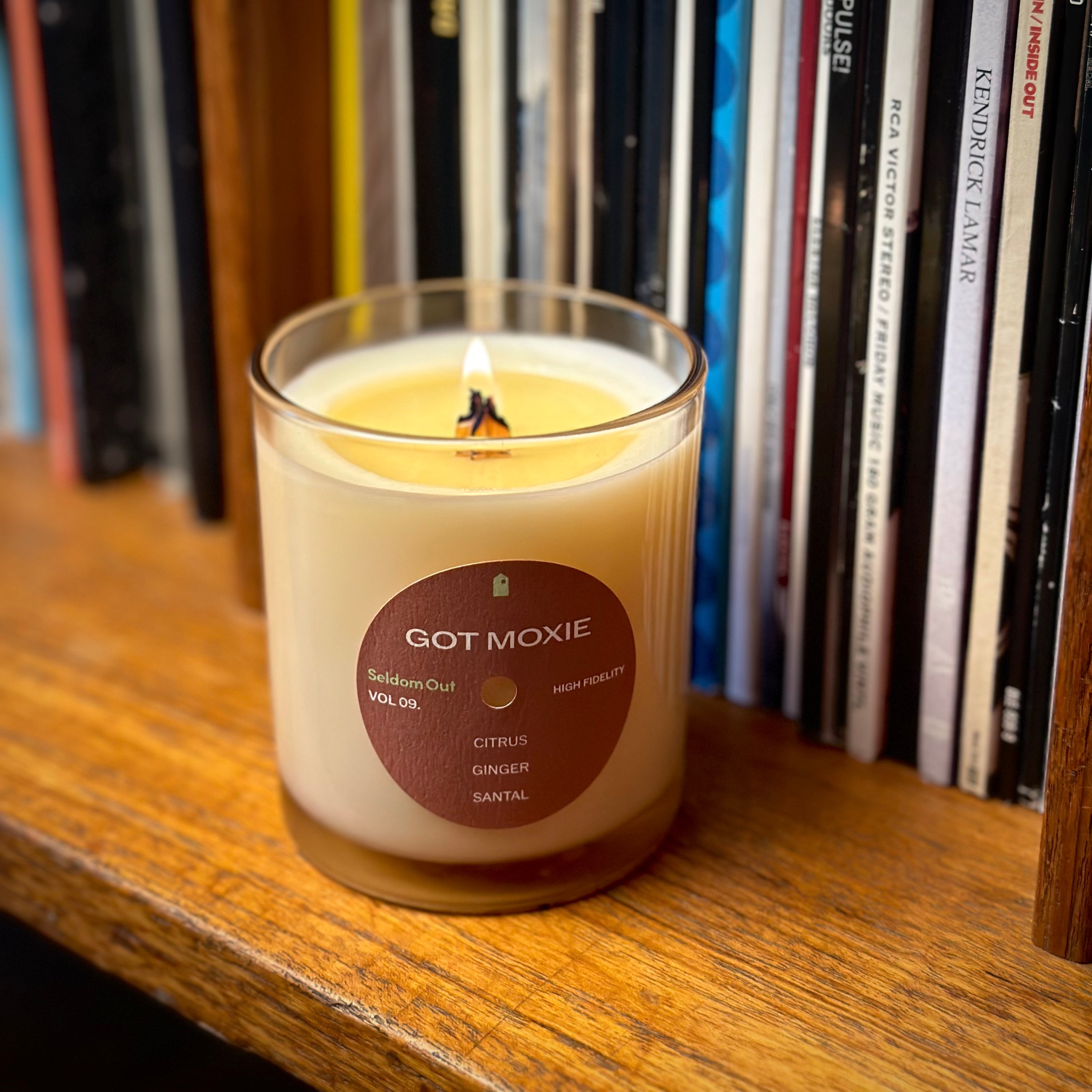 A lifestyle portrait image of our 8oz Got Moxie candle. The wick is alight. It has a slightly brown tinted glass vessel, a round label with a hole at the center, like a vinyl record. It’s sitting on a wooden shelf, with a row of vinyl records in the background. The lighting is warm.