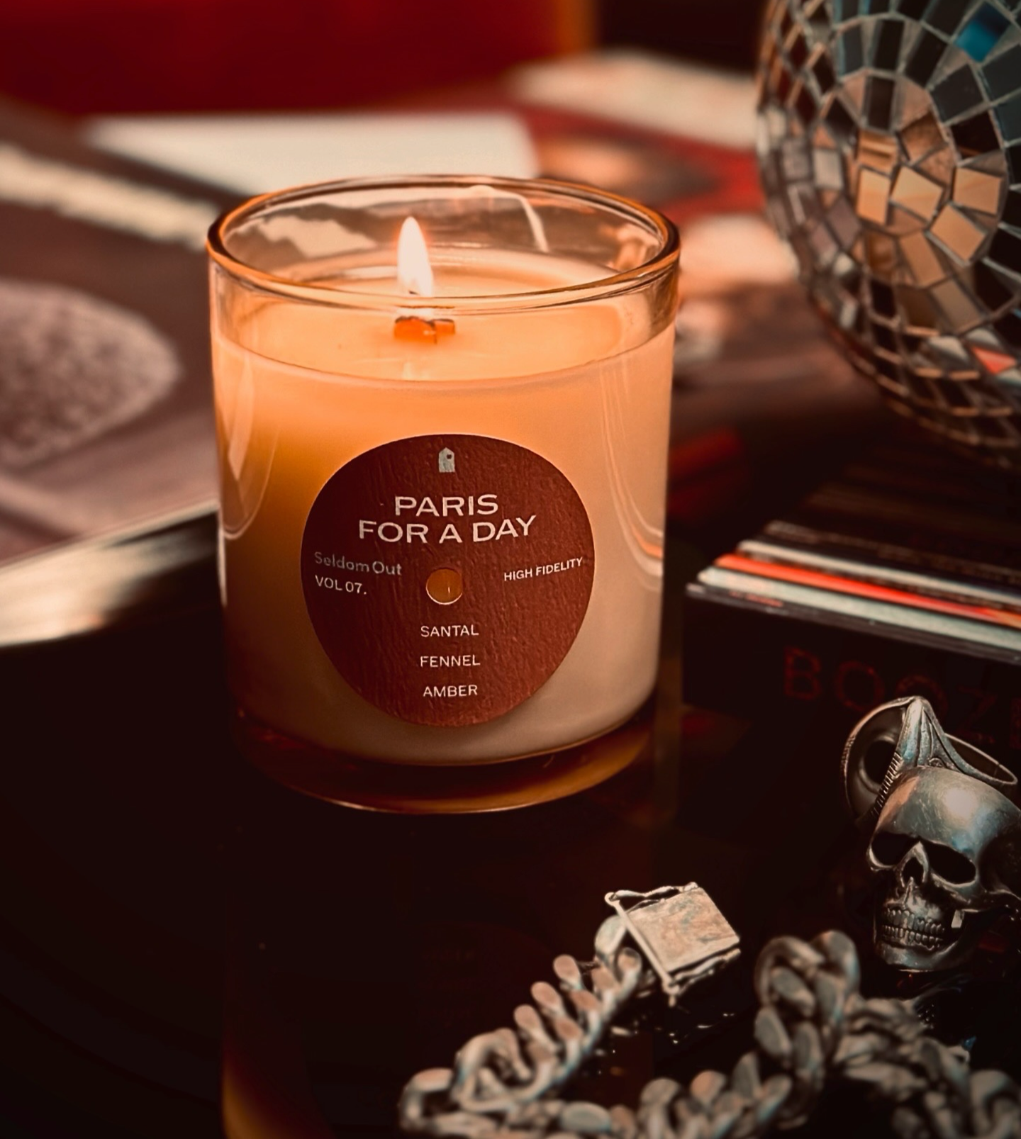 Our 8oz Paris for a Day candle sits on a dark glass table. The wooden wick is lit casting a warm glow. To one side sits a stack of vinyl records, in front some chunky silver jewelry and behind is a disco ball. The lighting is minimal, letting the candle throw moody shadows.