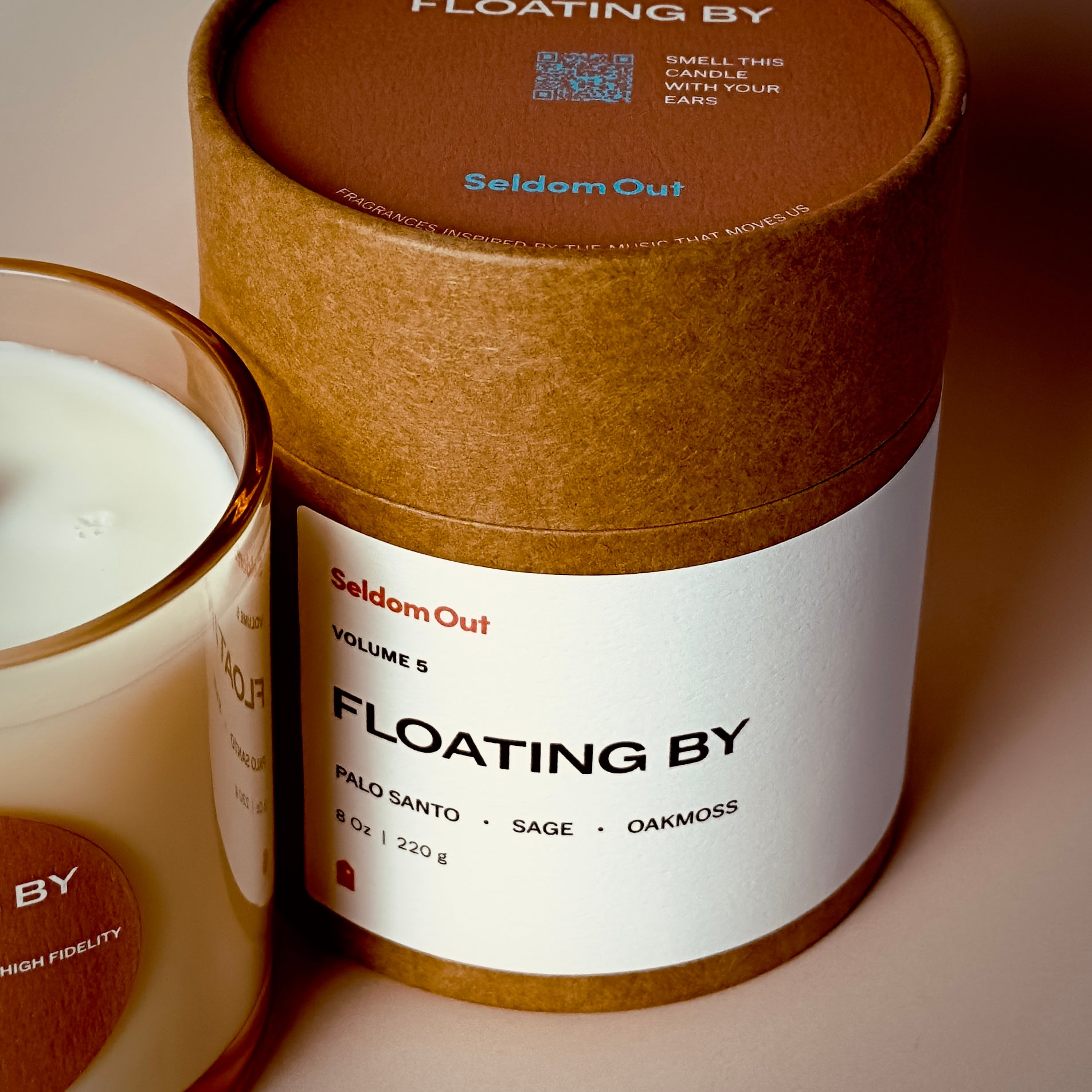A portrait image of our 8oz Floating By candle standing side by side with its packaging. The candle has a slightly brown tinted, straight sided glass vessel, a round label with a hole at the center, like a vinyl record. The Kraft tube packaging has a wide label on the front, with a round label on top. The label on top features a QR code with the instructions to “smell this candle with your ears”.
