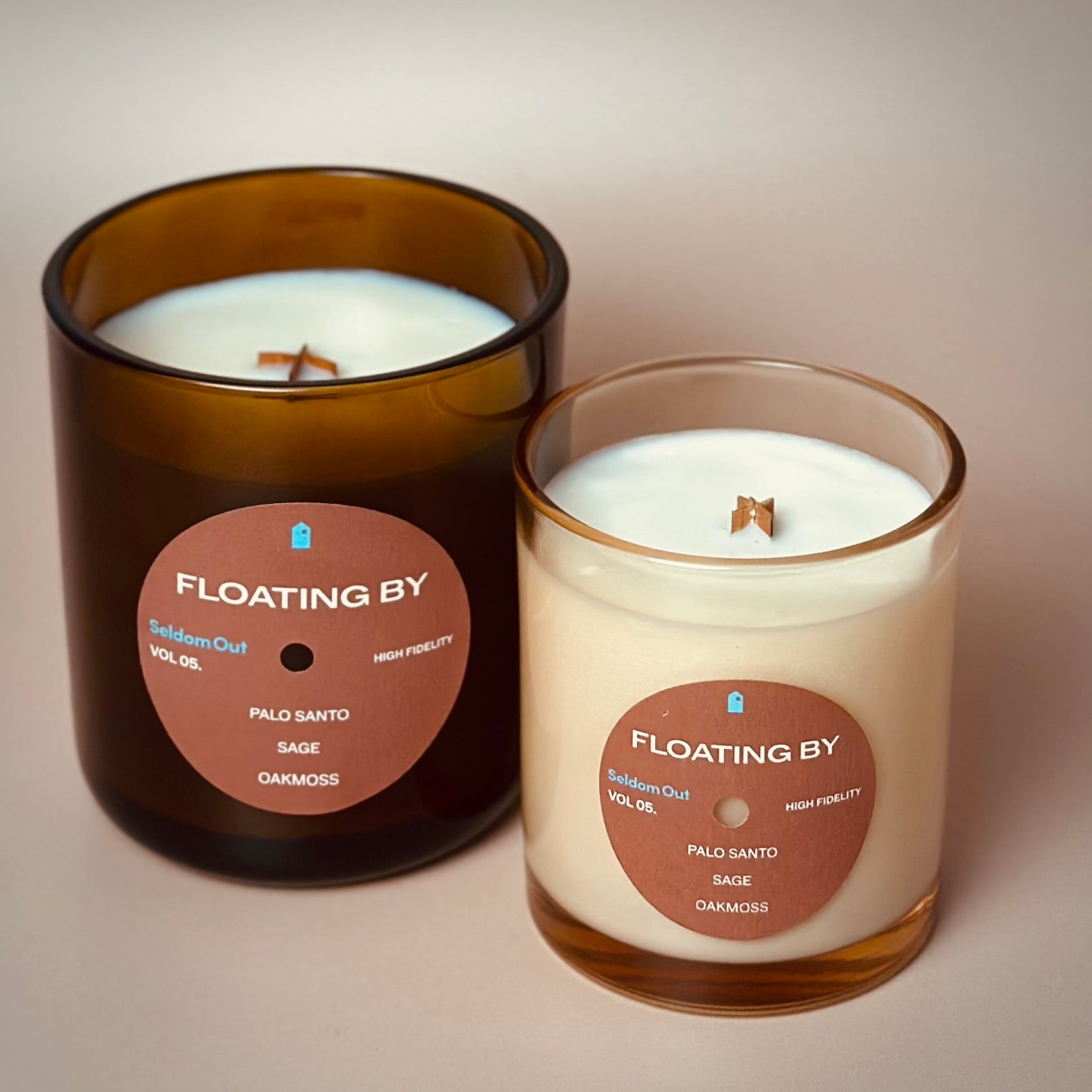 A portrait style image of our 8oz and 12oz Floating By Scented Candle standing side by side. The 8oz features a lightly tinted glass vessel, the 12 oz candle is in a heavy amber glass vessel. Both candles have a round front label.