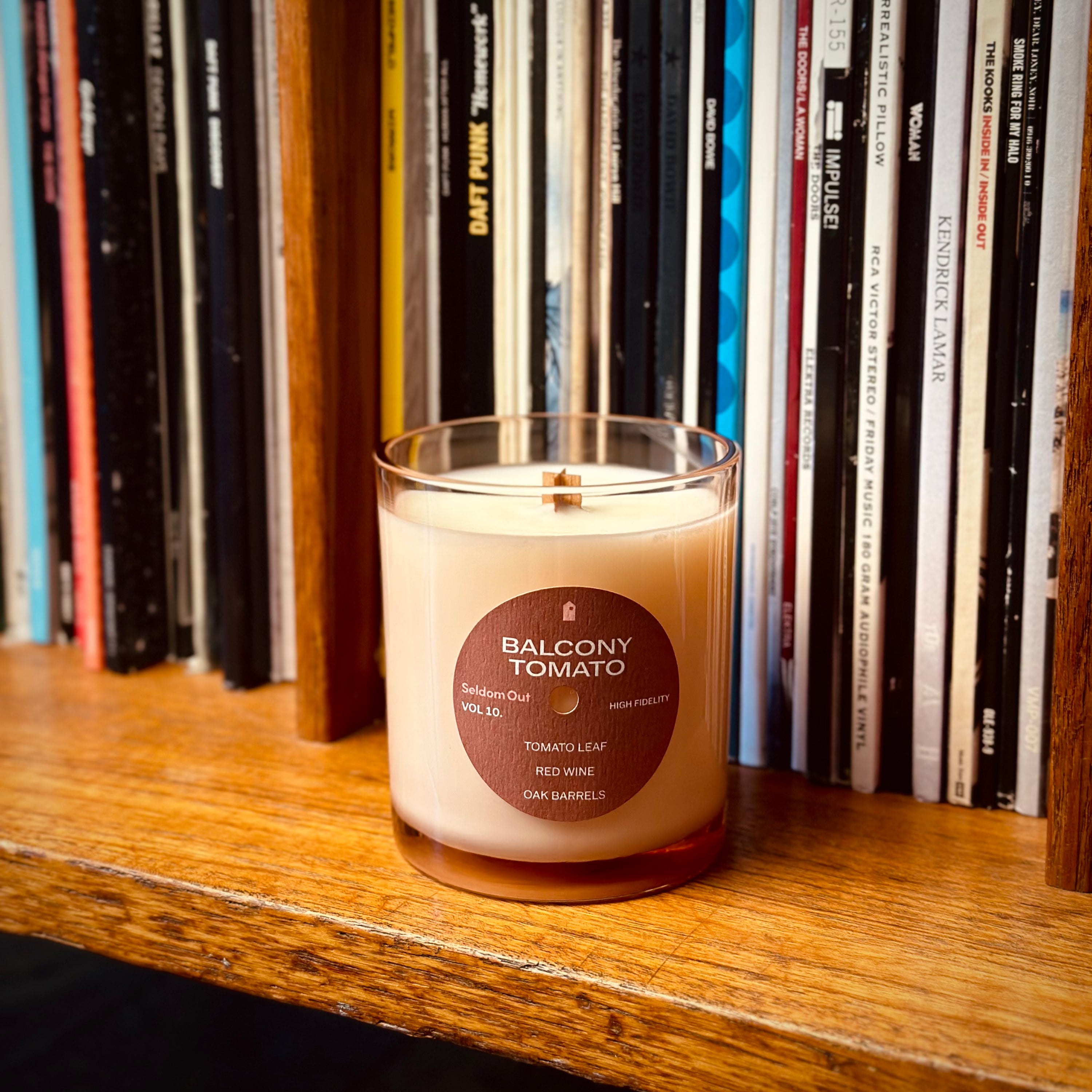 A lifestyle image of our 8 oz Balcony Tomato scented candle. It sits on a wooden shelf with a row of upright vinyl records in the background.