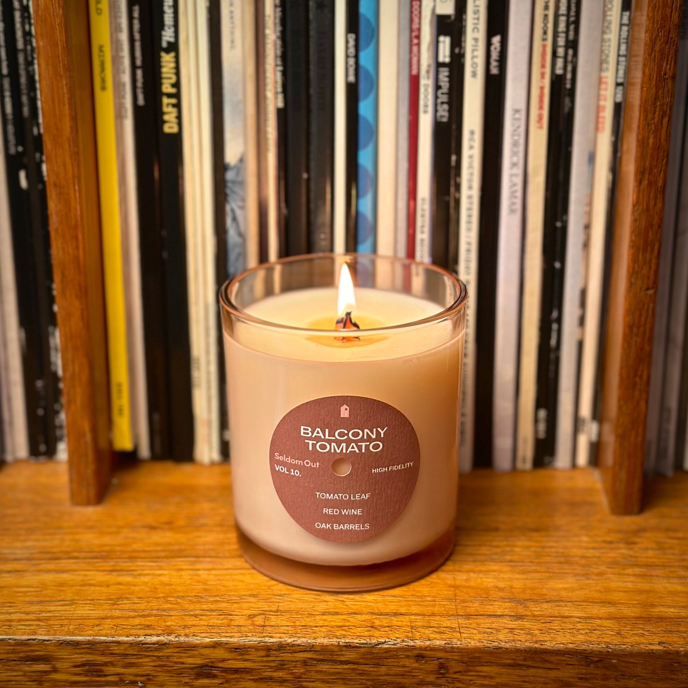 A portrait shot of our Balcony Tomato scented candle. A tinted glass vessel with a brown round label on the front. The label has a hole at the center making it look like a vinyl record. It is sitting on a wooden shelf in front of a row of vinyl records. The lighting is diffused and warm.