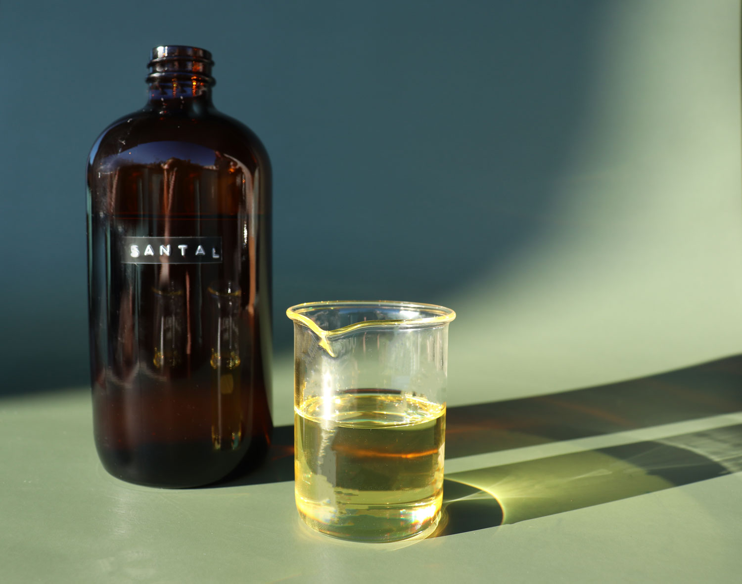 An amber glass bottle with no lid and labeled Santal stands behind a small glass science beaker half full of fragrance oil.