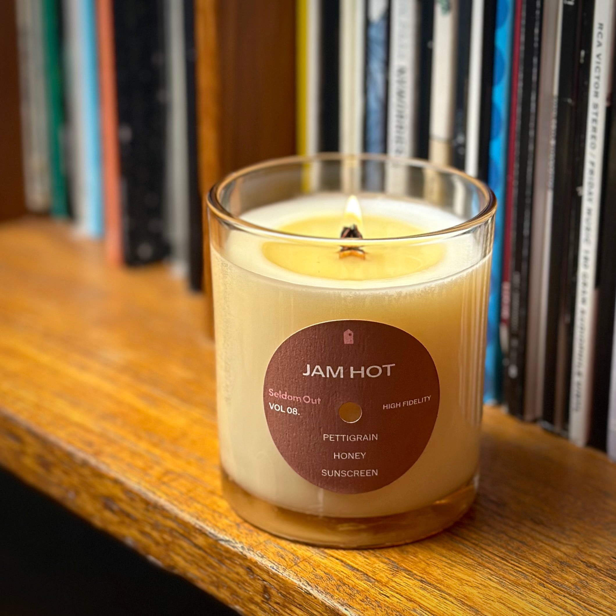 A lifestyle image of our 8oz Jam Hot scented candle. The wick is lit, it’s sitting on a wooden shelf with a row of vinyl records in the background.