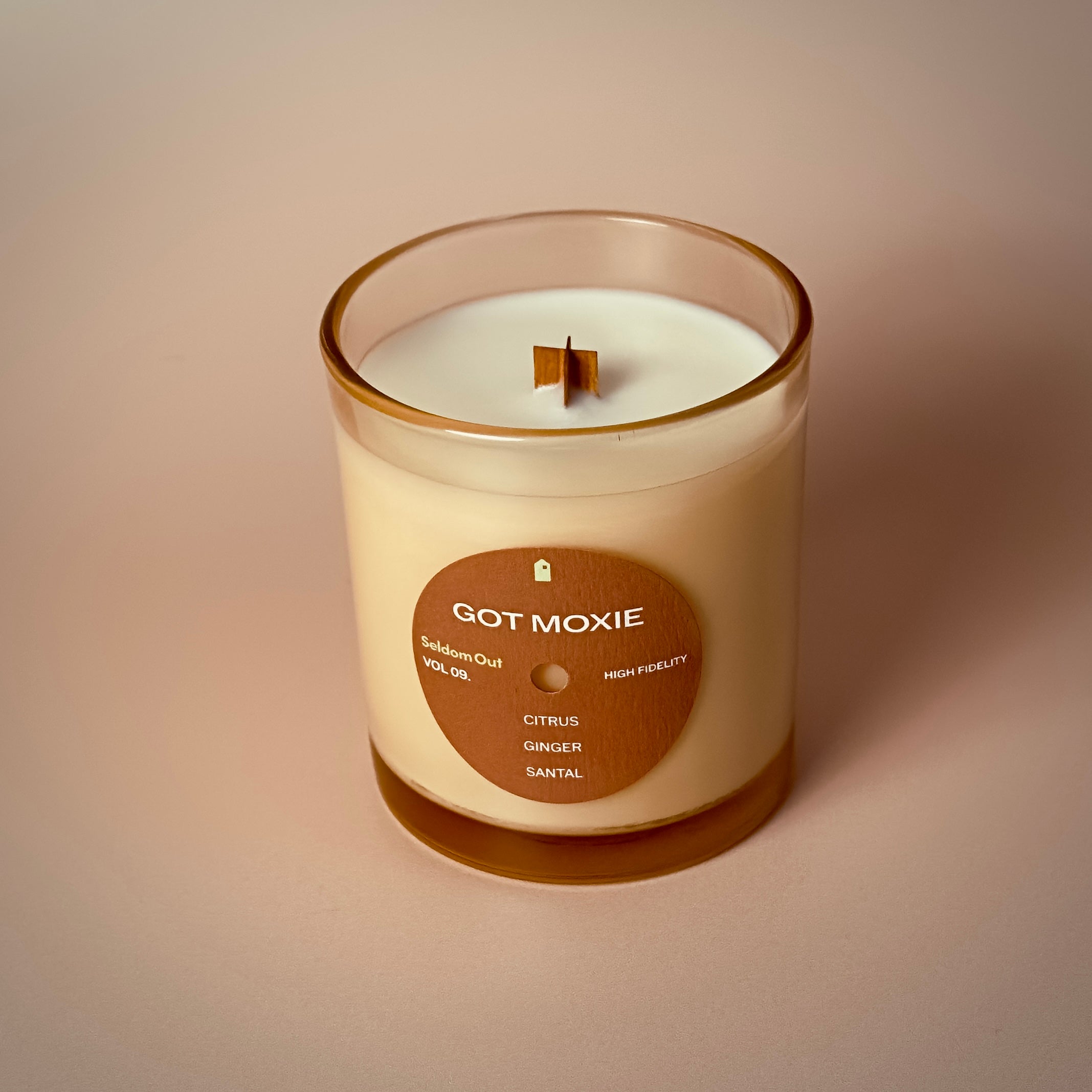 A portrait image of our 8oz Got Moxie candle. It has a slightly brown tinted glass vessel, a round label with a hole at the center, like a vinyl record.