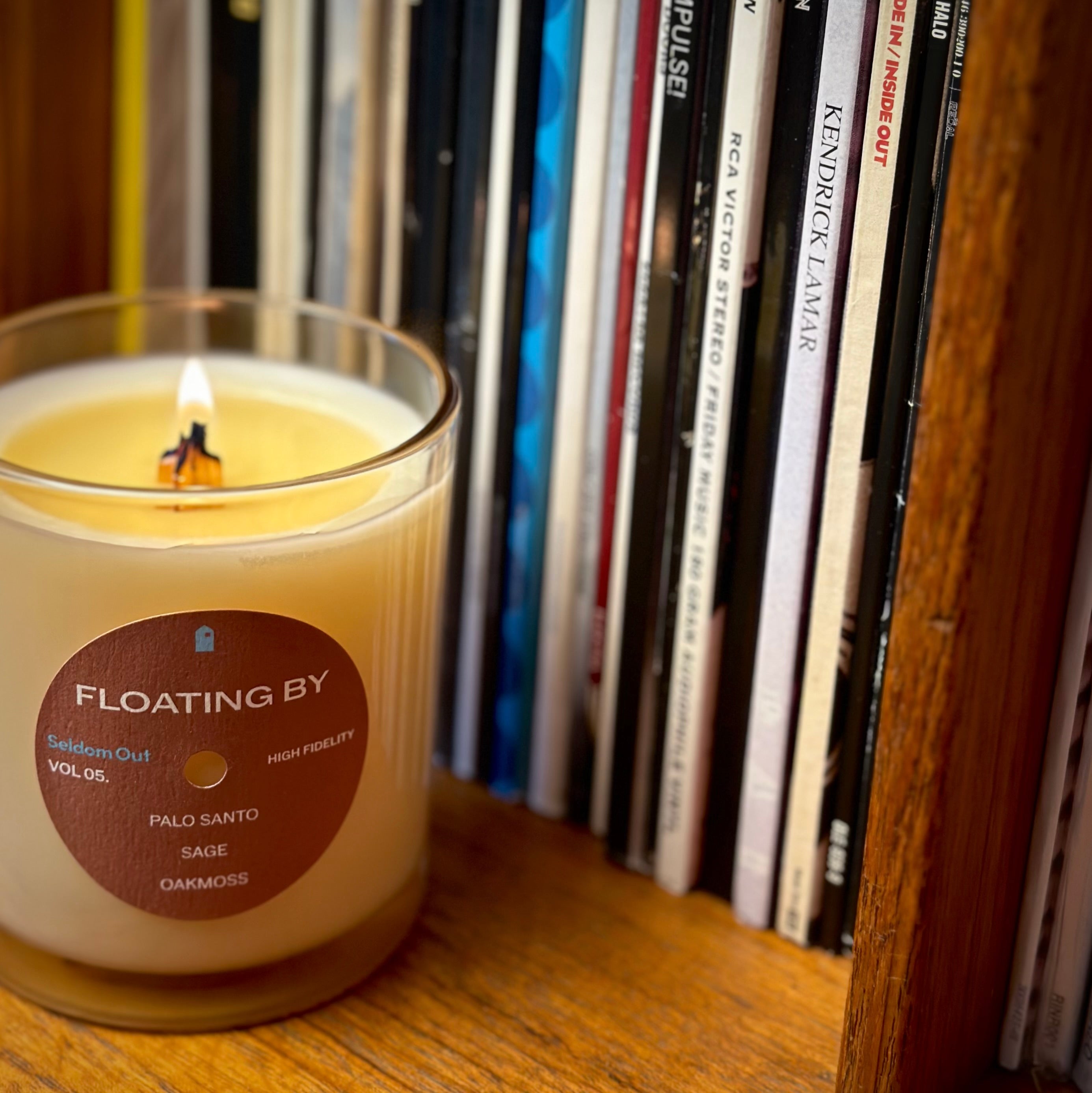 A portrait image of our 8oz Floating By candle. It has a slightly brown tinted glass vessel, a round label with a hole at the center, like a vinyl record. The wick is lit with a tall cylindrical flame, it’s sitting on a wooden shelf with a row of vinyl records in the background.