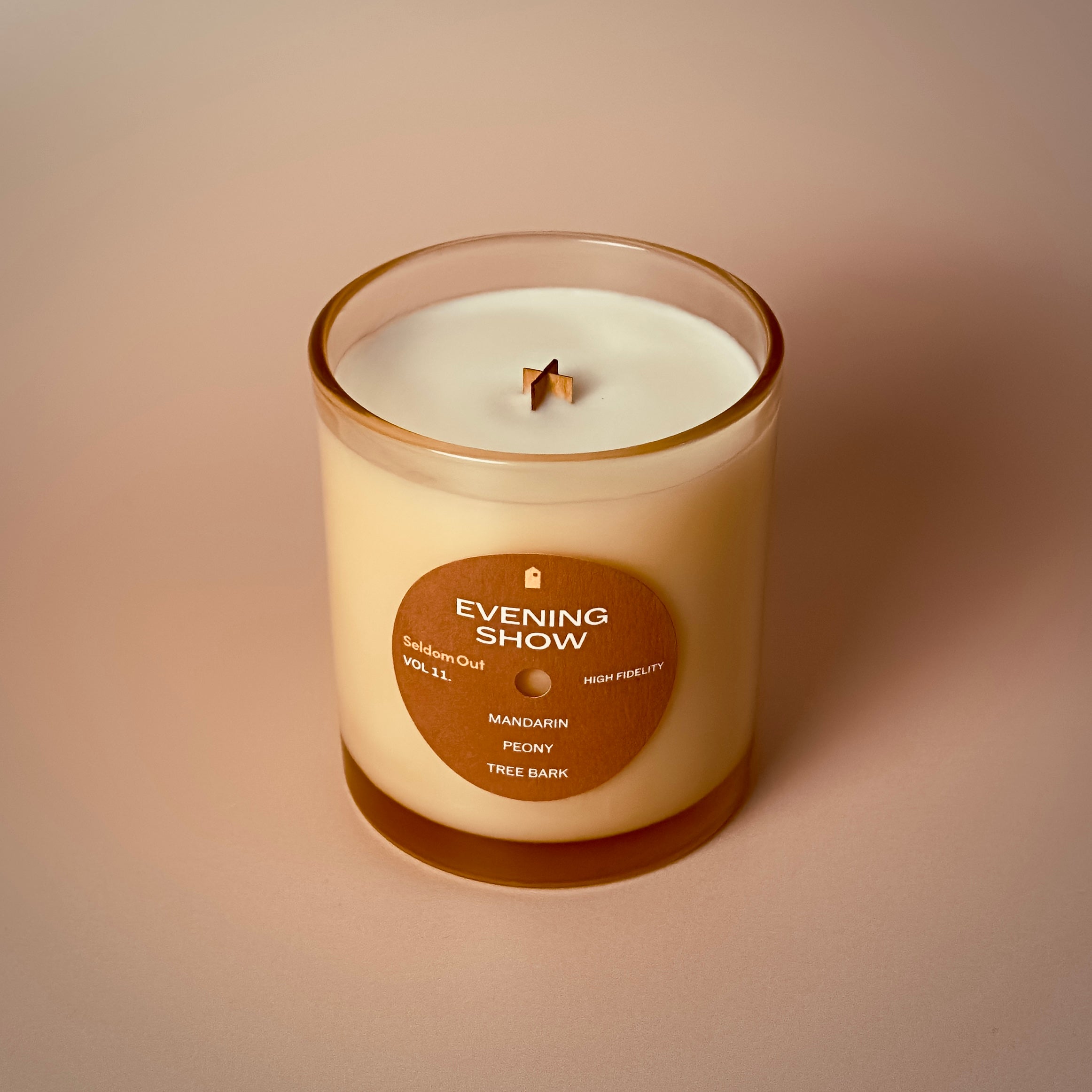 A portrait image of our 8oz Evening Show candle. It has a slightly brown tinted glass vessel, a round label with a hole at the center, like a vinyl record.