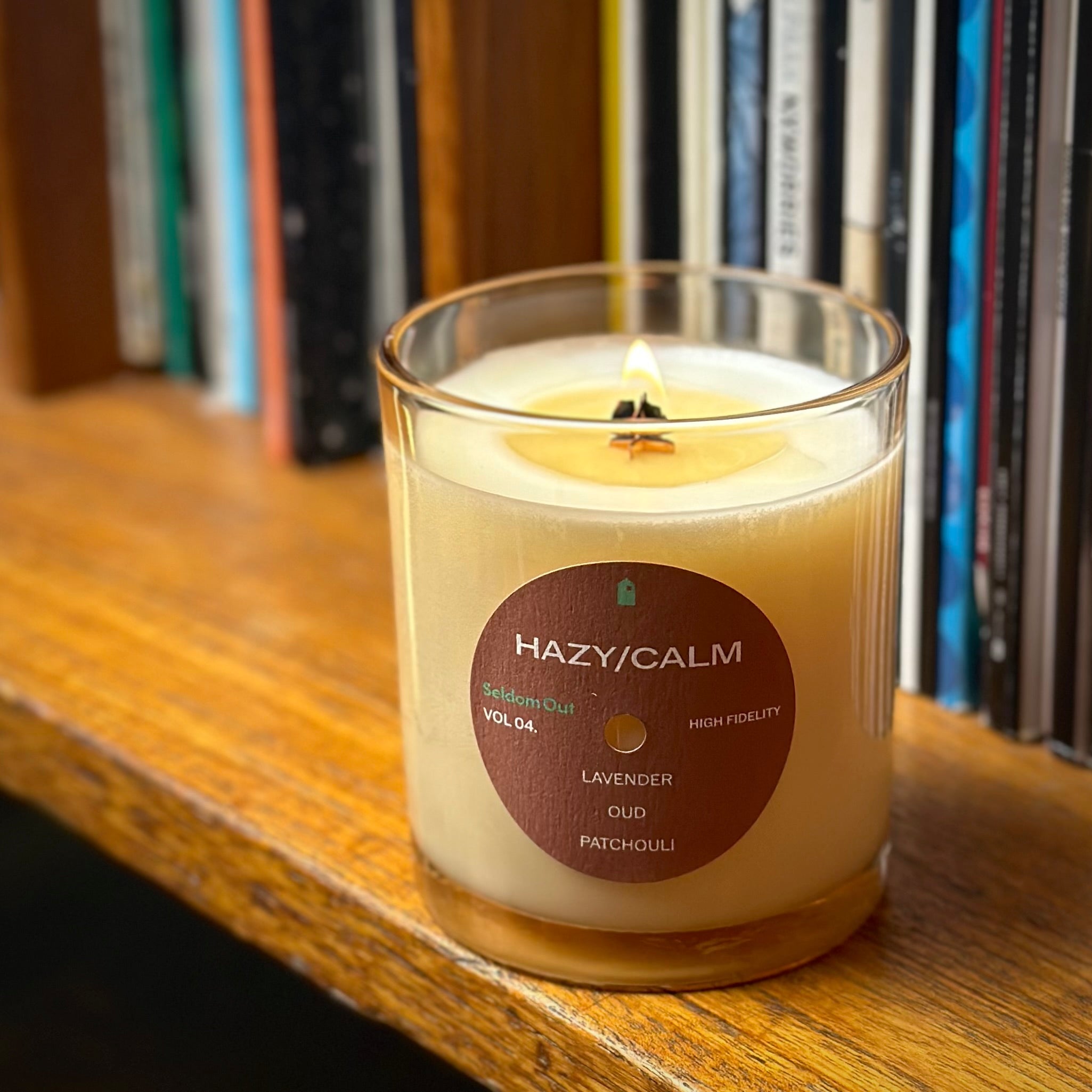 A lifestyle portrait image of our 8oz Hazy / Calm scented candle. The wick is alight, with a cylindrical flame. It has a slightly brown tinted glass vessel, a round label with a hole at the center, like a vinyl record. It sits on a wooden shelf, with rows of vinyl records in the background.