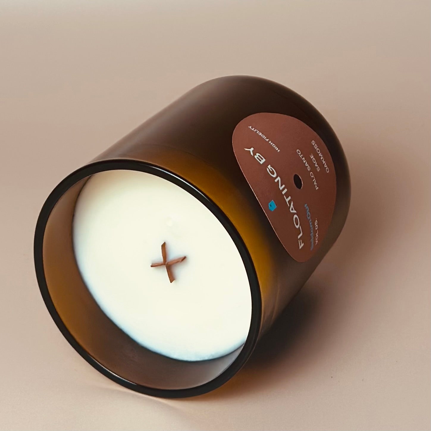 A portrait image of our 12oz  Dreamwave candle, laying on it’s side to highlight the wooden x shaped wick. The candle has a heavy amber glass vessel, a round label with a hole at the center, like a vinyl record.