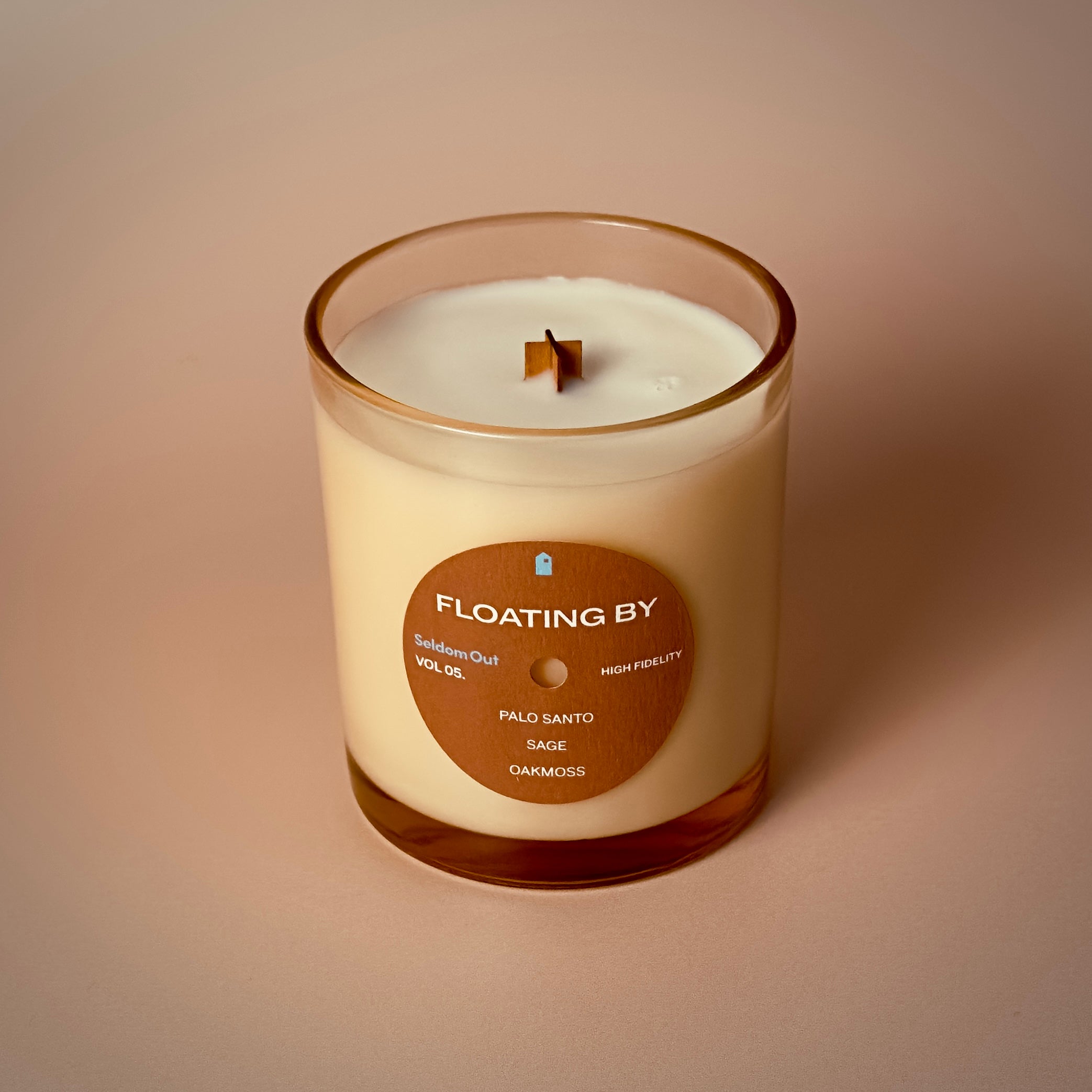 A portrait image of our 8oz Floating By candle. It has a slightly brown tinted glass vessel, a round label with a hole at the center, like a vinyl record.