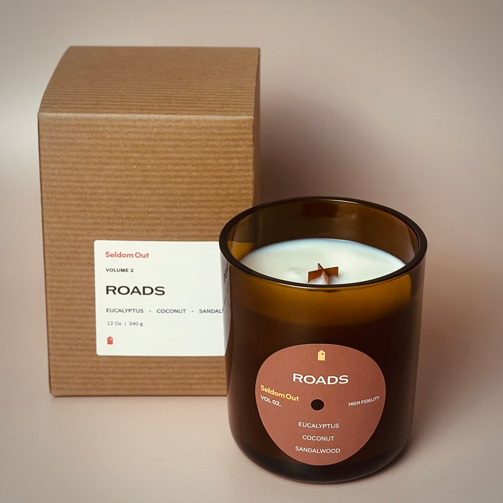 A portrait image of our 12oz Roads scented candle made in a heavy amber glass jar, standing next to its candle box. The candle features a round label, with a hole in the middle, that sort of looks like a vinyl record. The box is made of Kraft paper with a rectangle label, that has been folded to go around a corner.