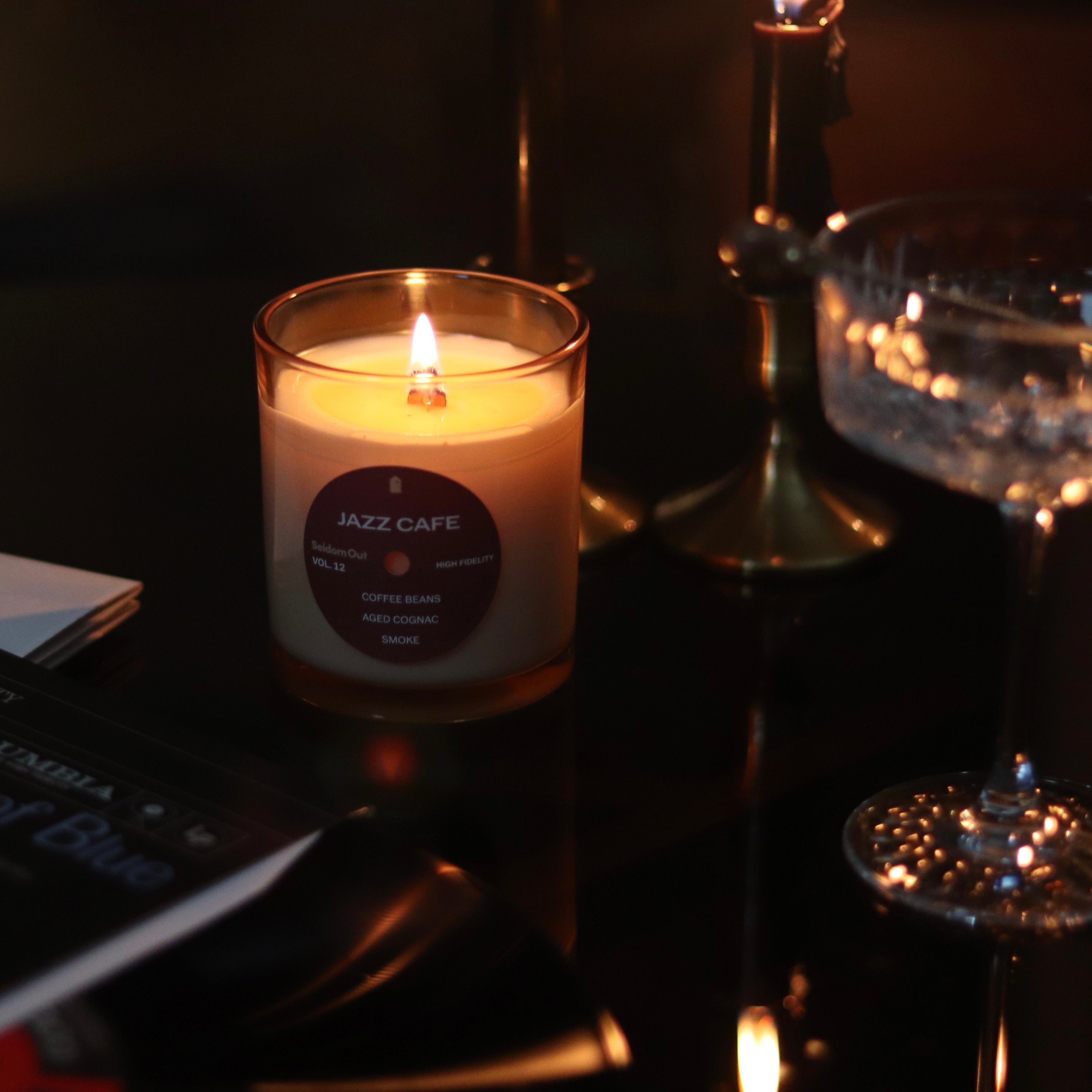 A lit 8oz Jazz Cafe candle glows warmly in a dark, intimate setting. The candle's wooden wick casts a gentle light across its cream-colored wax and burgundy label. In the shadowy background, martini glasses sparkle and dark shapes suggest vinyl records or books, creating an atmosphere reminiscent of a late-night jazz lounge.