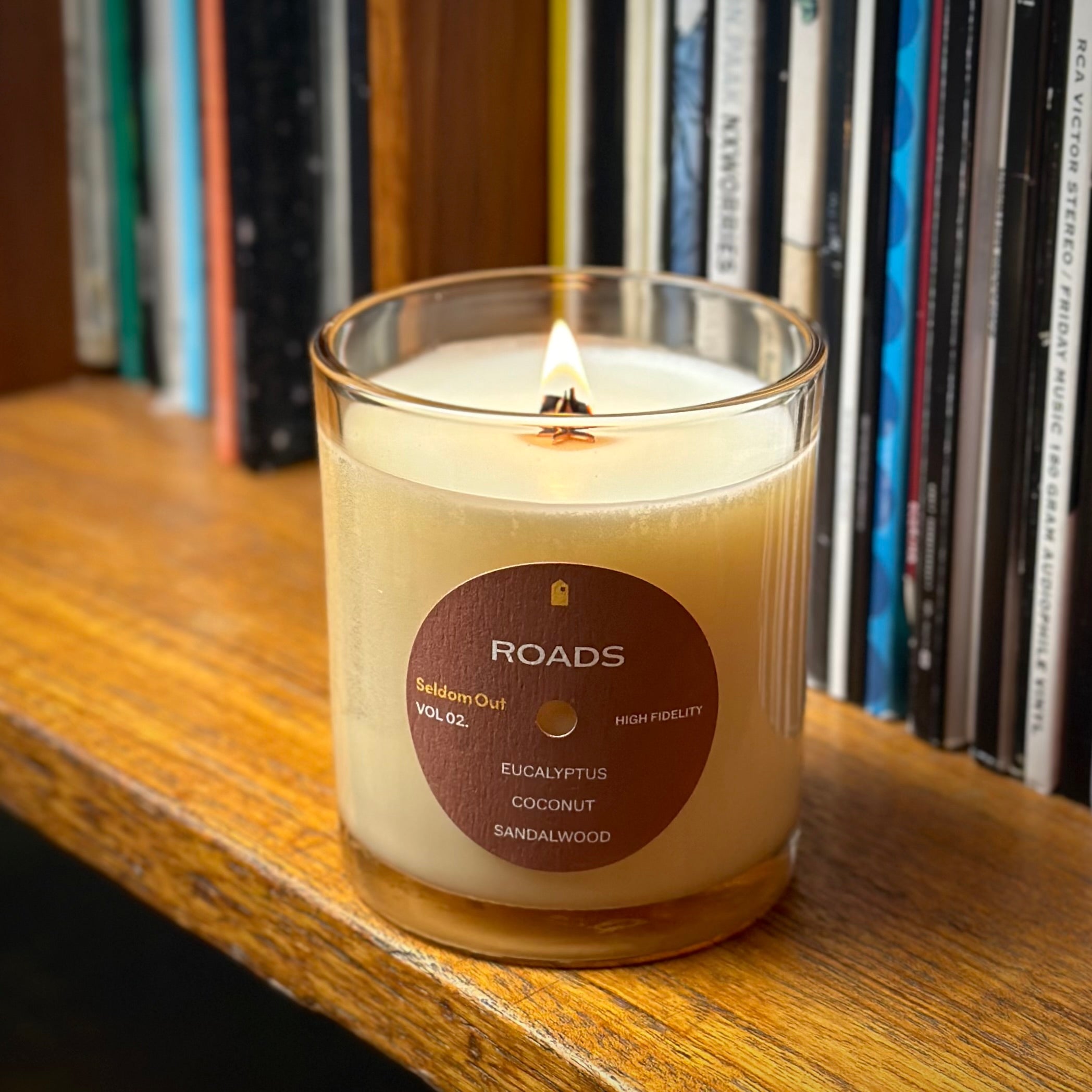 A lifestyle image of our 8oz Roads scented candle. It features a round label, with a hole in the middle, that sort of looks like a vinyl record. The wick is lit and it sits on a wooden shelf, in the background stacks of vinyl are visible.