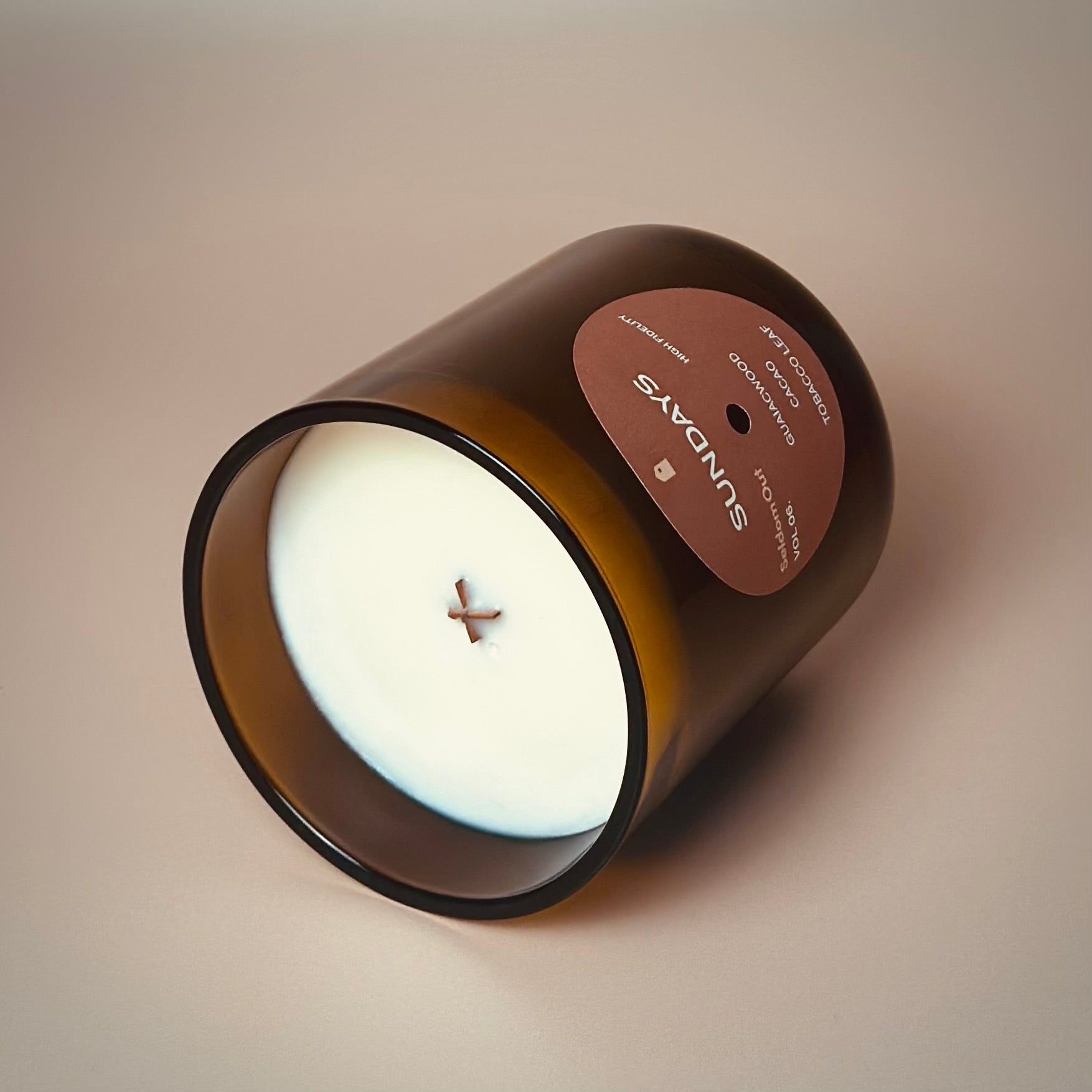 A portrait image of our 8oz Sundays scented candle made in a light amber tinted glass jar. It features a round front label, that has a hole cut out in the middle, not unlike a vinyl record. The candle is laying on its side to highlight the wooden x shaped wick.