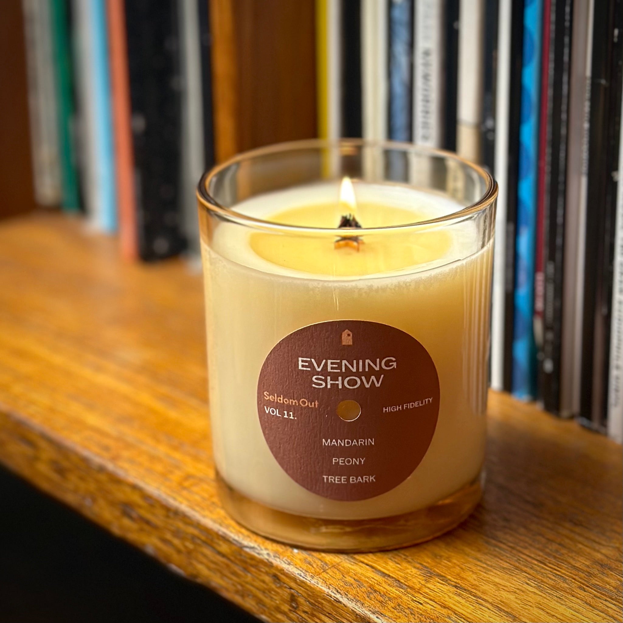 A portrait image of our 8oz Evening Show candle with its wick alight. It has a slightly brown tinted glass vessel, a round label with a hole at the center, like a vinyl record. It’s sitting on a wooden shelf, in the background is a row of vinyl records.