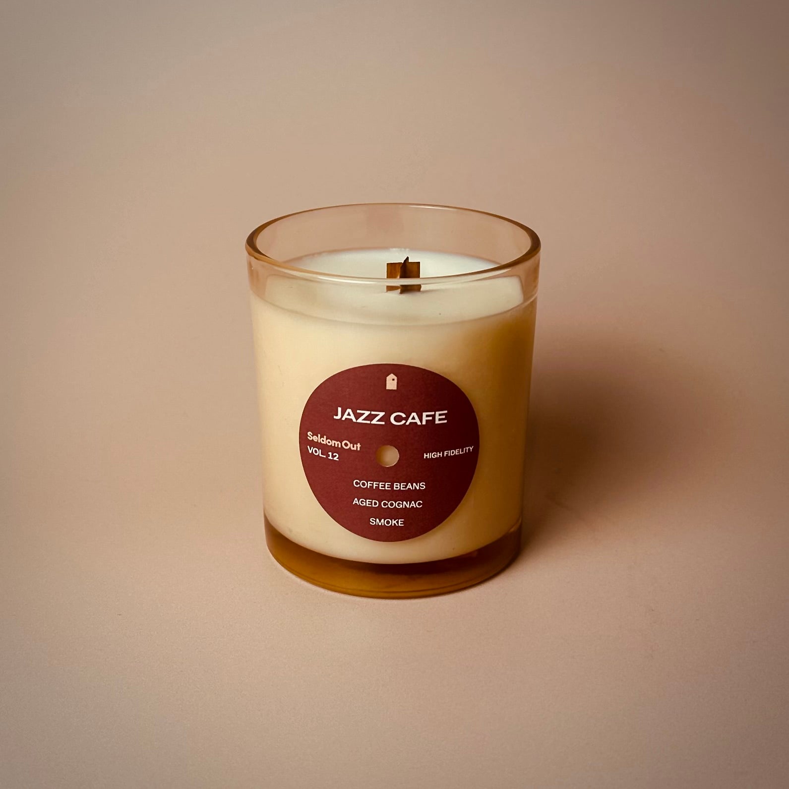 A cream-colored scented candle in a clear glass vessel with a wooden wick. The candle features a circular burgundy label designed to look like a vinyl record label, reading 'JAZZ CAFE' with 'Seldom Out VOL-12' and listing three scent notes: Coffee Beans, Aged Cognac, and Smoke. The candle is photographed against a neutral beige background.