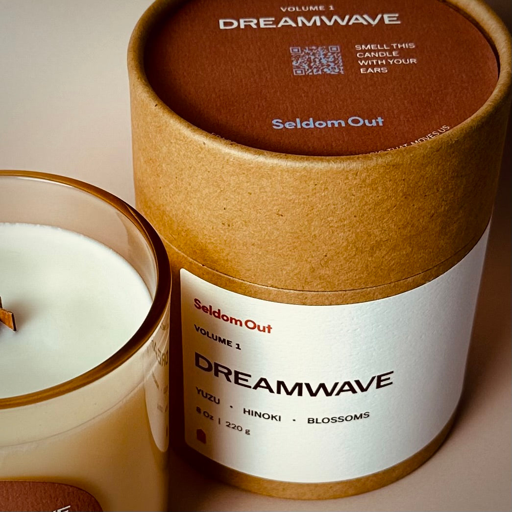 A portrait image of our 8oz Dreamwave candle packaging, alongside the candle. The Kraft Tube packaging features a wide label on the front with an additional label on the top showing a QR Code & the instructions to smell this candle with your ears.