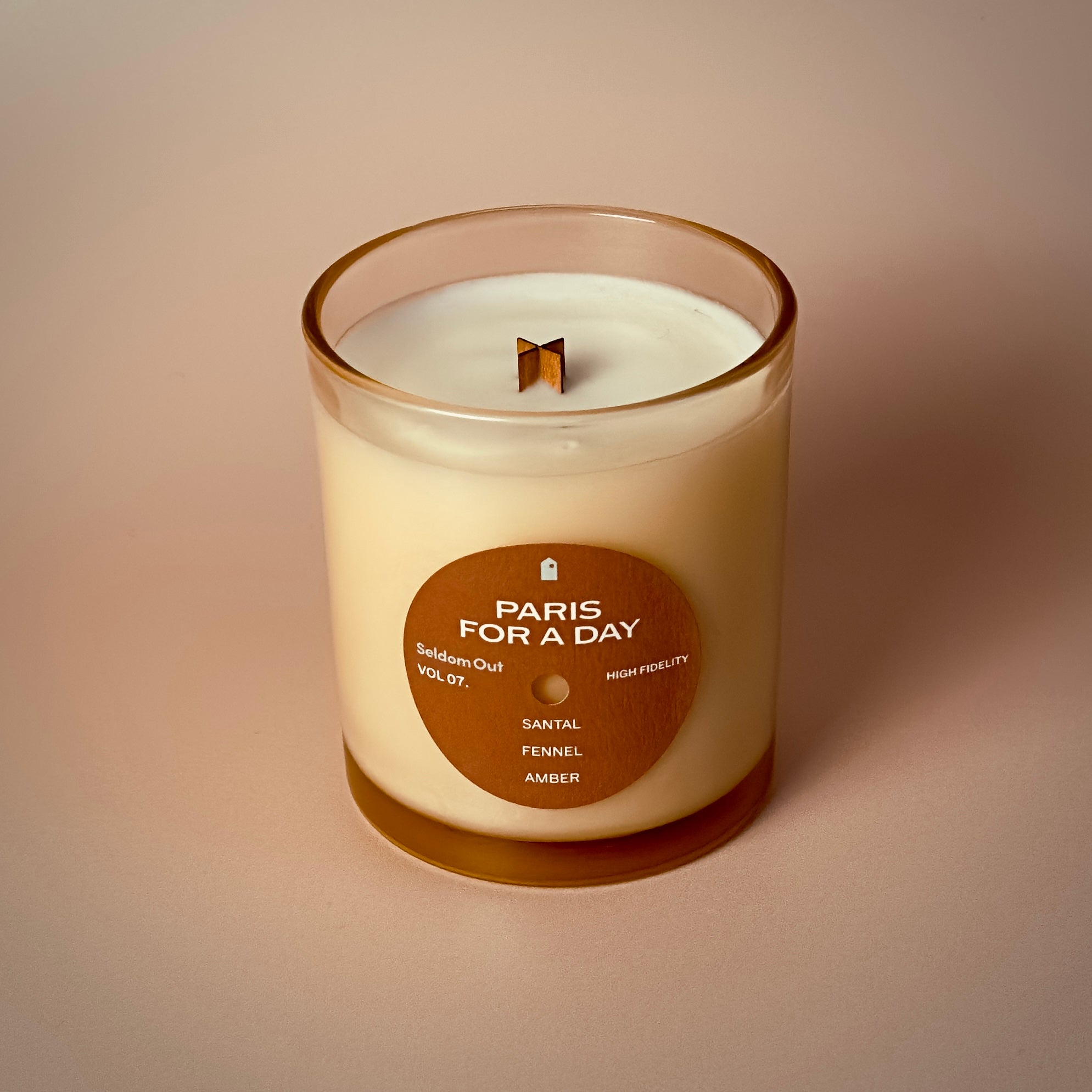 A portrait image of our 8oz Paris For a Day scented candle. It features a round label, with a hole in the middle, that gives the vibe of a vinyl record. The lighting is warm & diffused with exaggerated shadows.