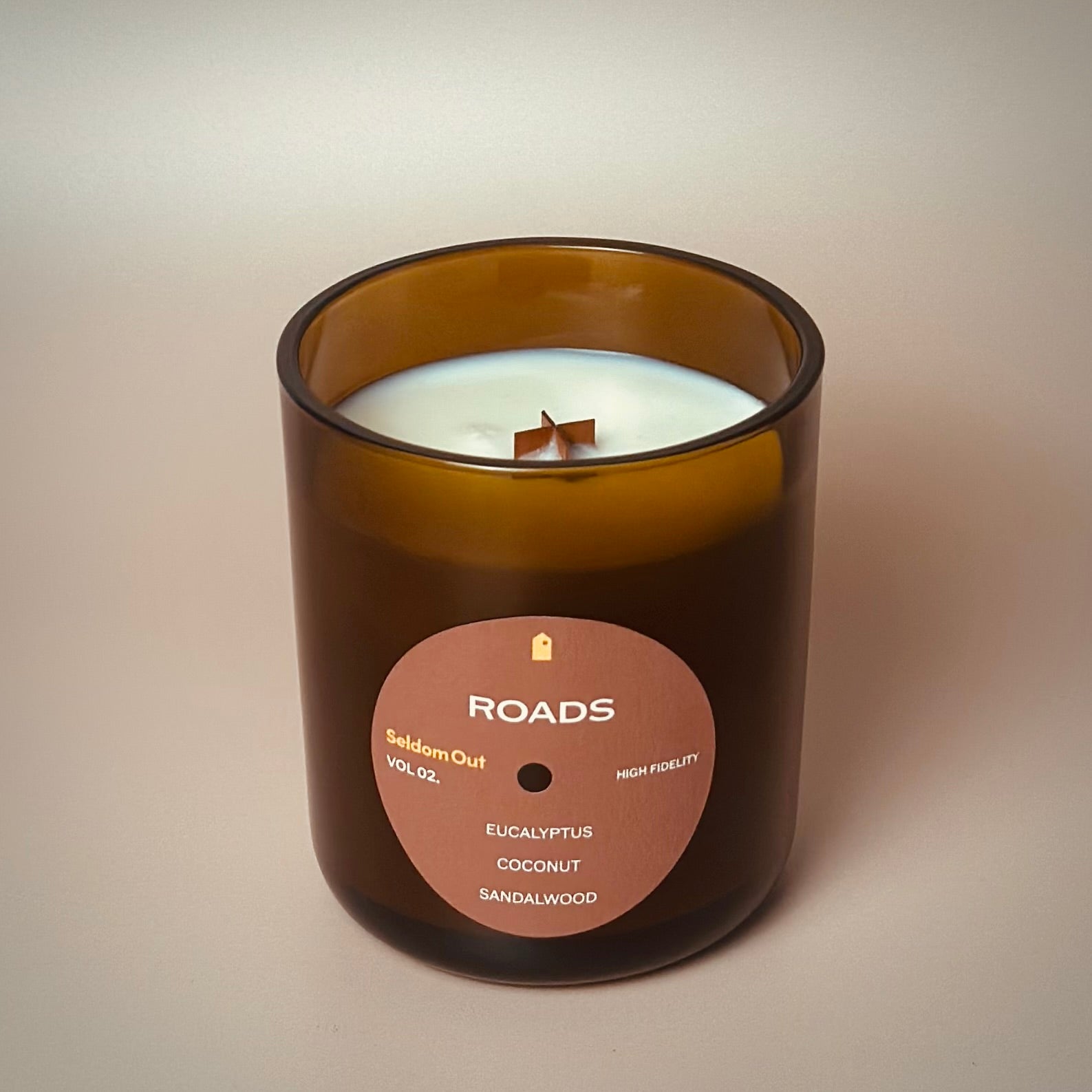 A portrait image of our 12oz Roads scented candle made in a heavy amber glass jar. It features a round label, with a hole in the middle, that sort of looks like a vinyl record.