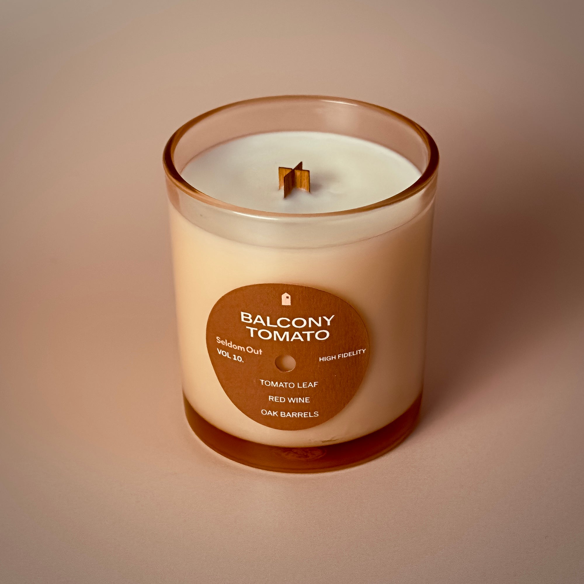 A portrait shot of our Balcony Tomato scented candle. A tinted glass vessel with a brown round label on the front. The label has a hole at the center making it look like a vinyl record. The lighting is diffused and warm.