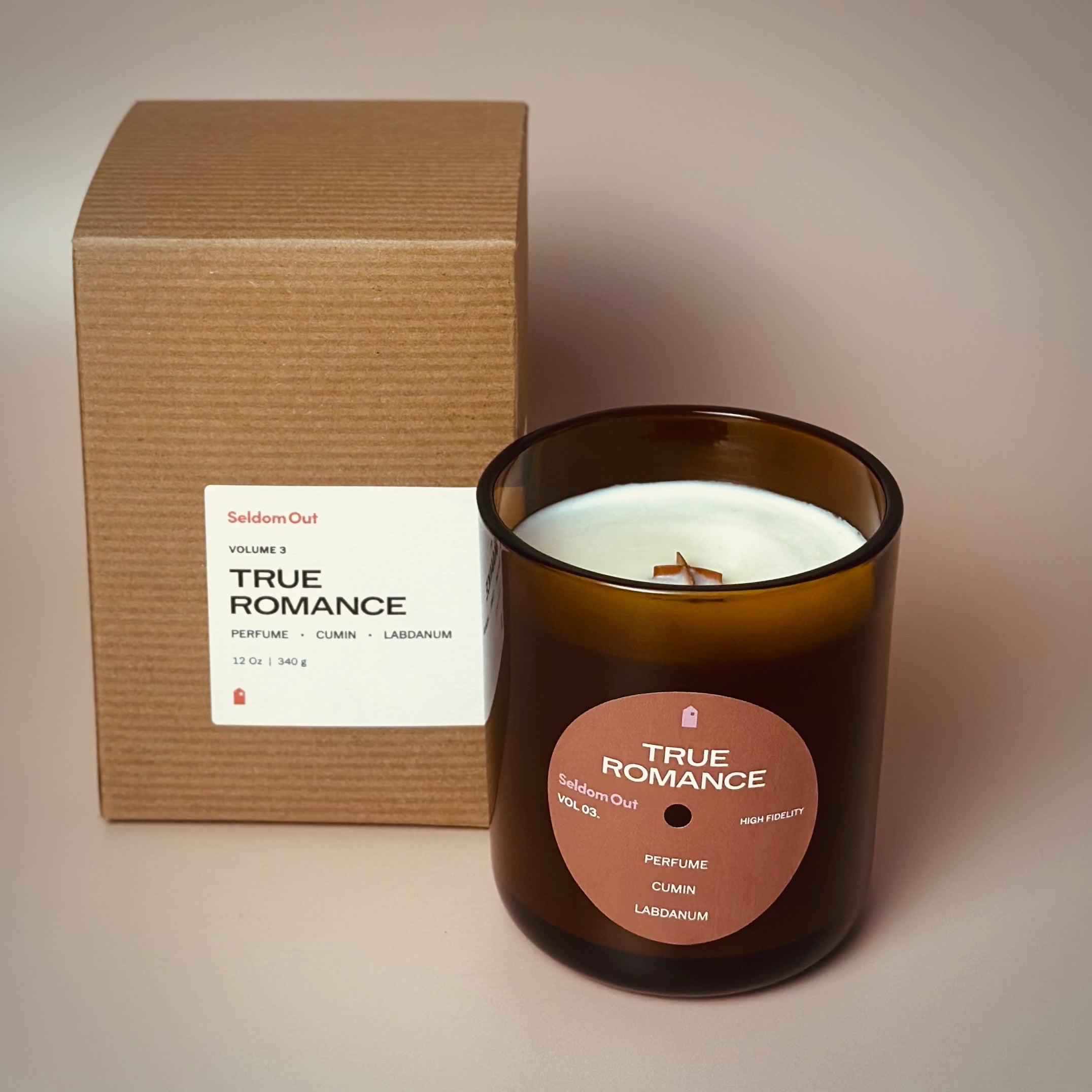 A portrait image of our 12oz True Romance scented candle made in a heavy amber glass jar, standing in front of a Kraft box. The candle features a round front label, that has a hole cut out in the middle, not unlike a vinyl record. The box has a wide rectangle label that wraps around one corner.