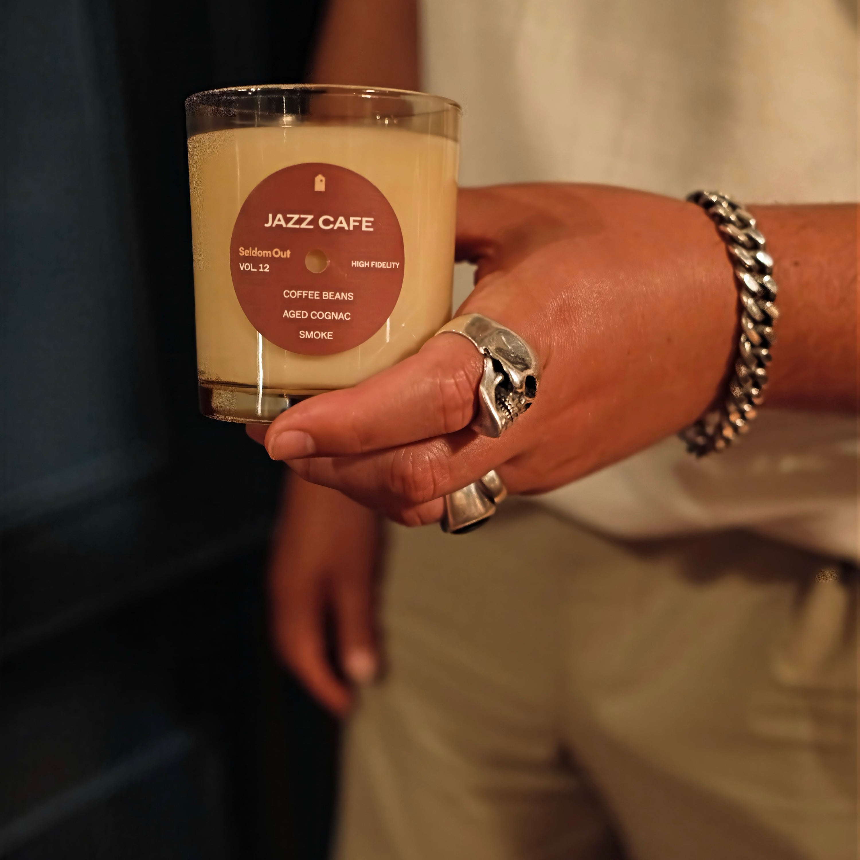 A close up image of a mans hand holding our 8oz Jazz Cafe scented candle. He is wearing heavy, chunky silver jewelry and the lighting is warm and moody.