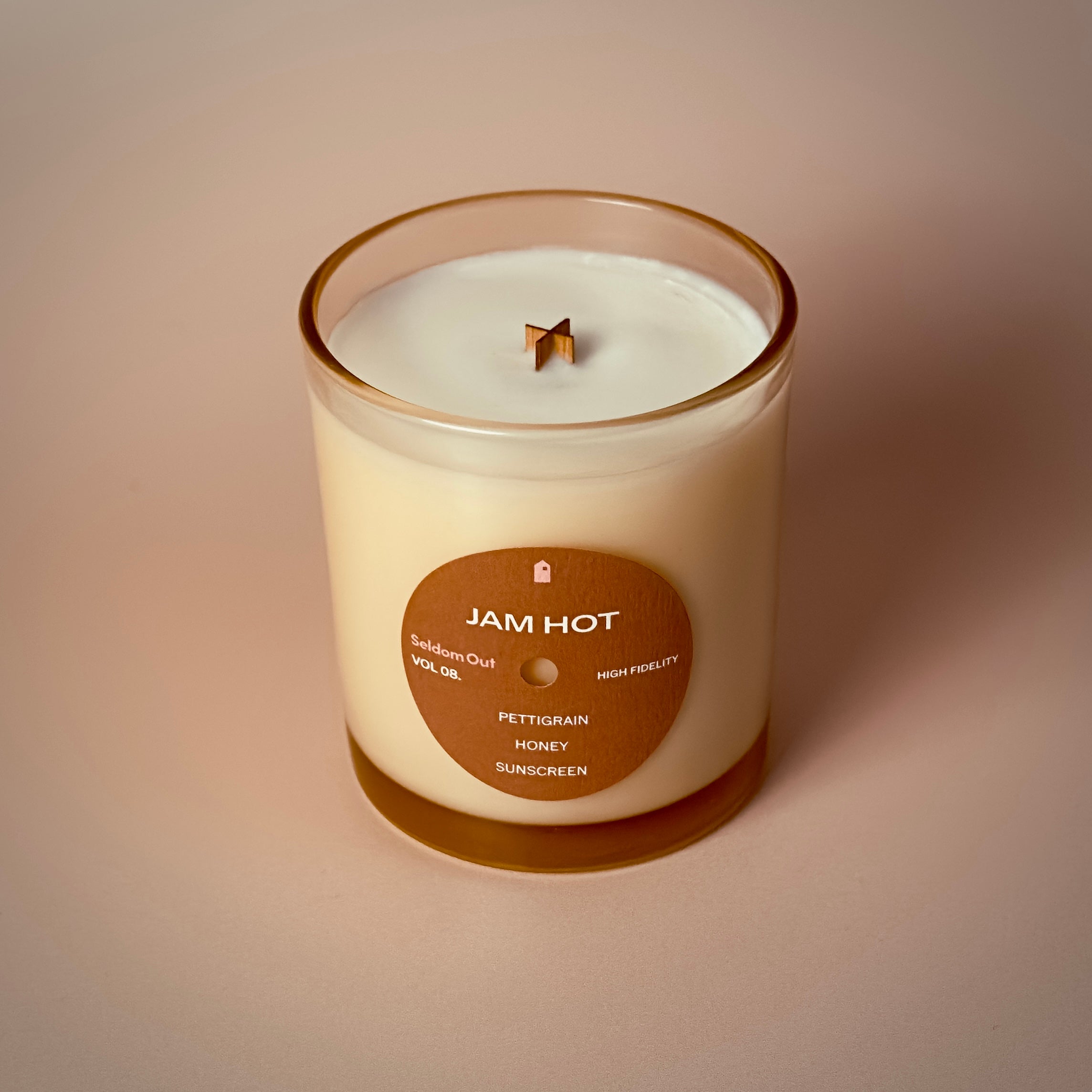 A portrait image of our 8oz Jam Hot scented candle. It features a round label, with a hole in the middle, that gives the vibe of a vinyl record. The lighting is warm & diffused with exaggerated shadows.