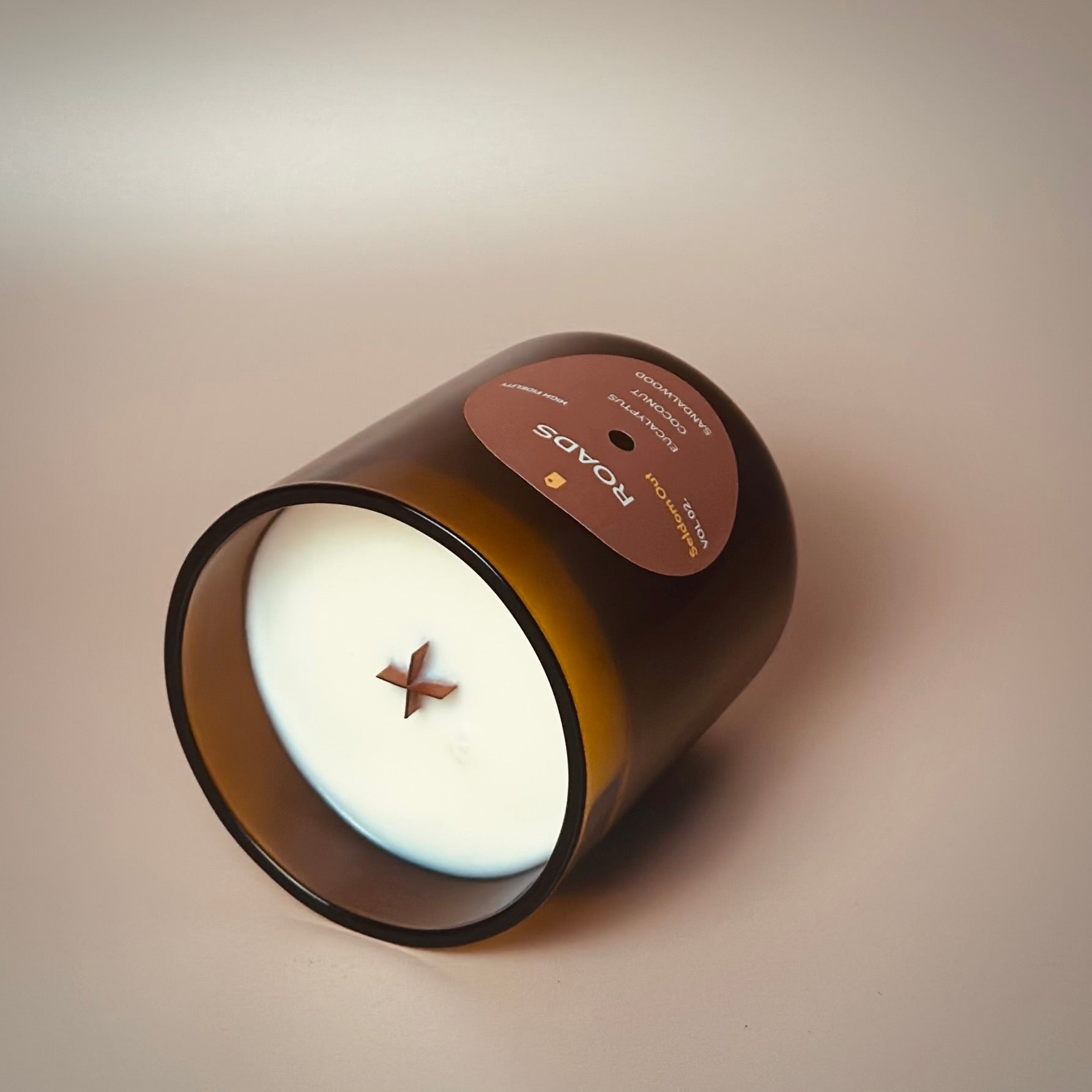 A portrait image of our 12oz Roads scented candle made in a heavy amber glass jar. It features a round label, with a hole in the middle, that sort of looks like a vinyl record. The candle is laying on its side to highlight the X shaped wooden wick.