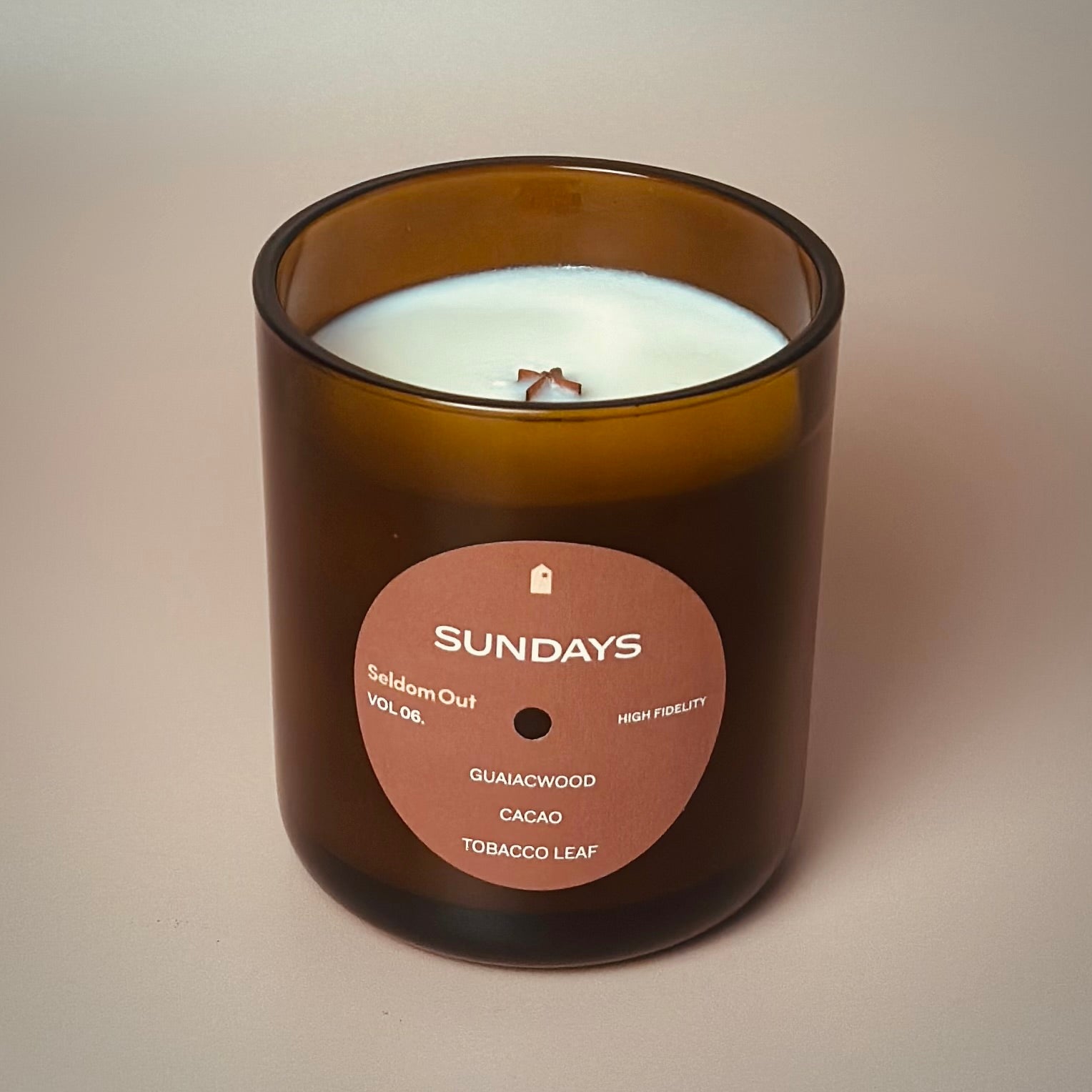 A portrait image of our 12oz Sundays scented candle made in a heavy amber tinted glass jar. It features a round front label, that has a hole cut out in the middle, not unlike a vinyl record.