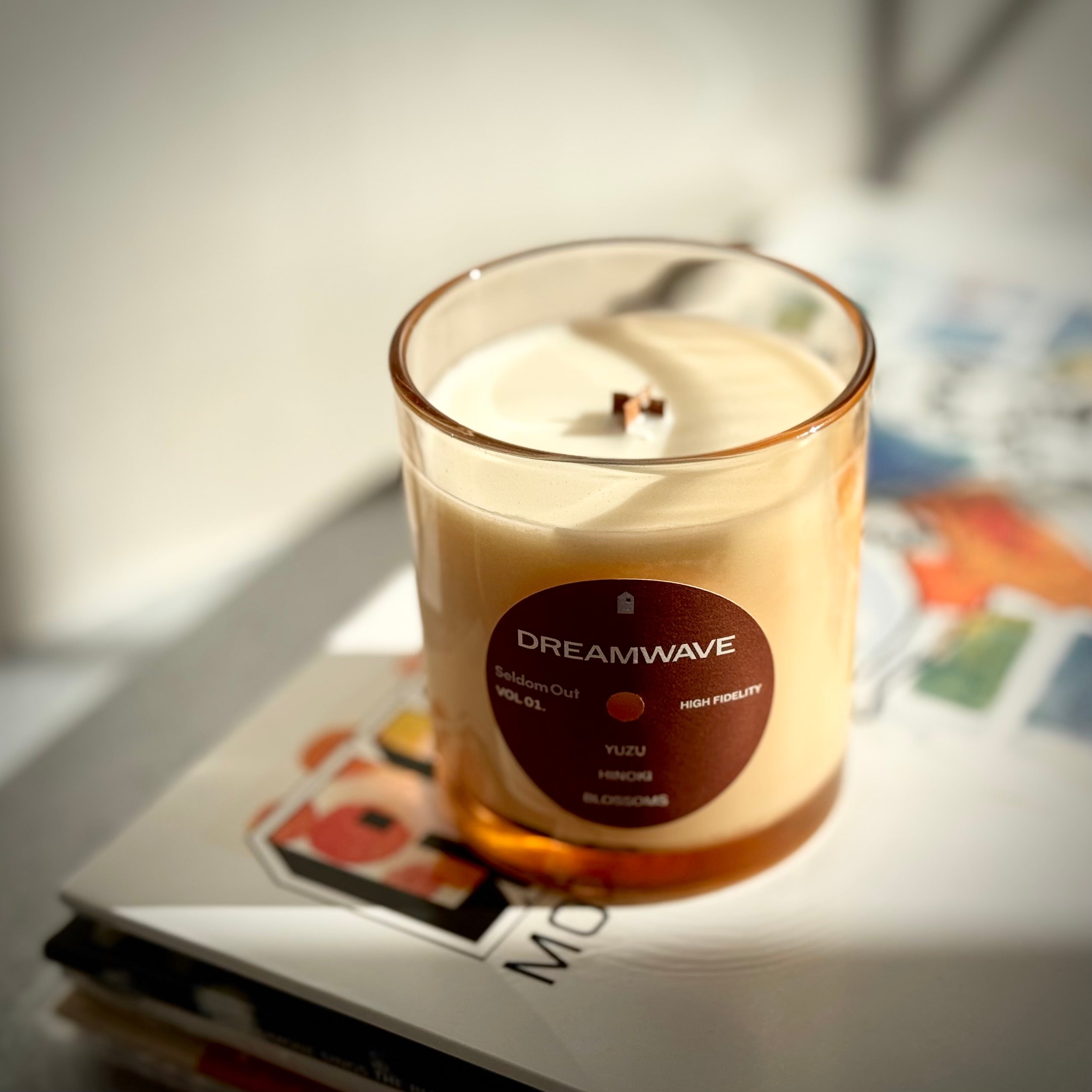 Portrait image of our 8oz Dreamwave Candle highlighting the vinyl record shaped label, tinted glass vessel and wooden x shaped wick. The candle sits in a bright scene with dramatic shadows on top of a stack of vinyl records.