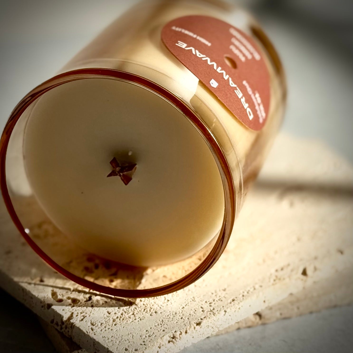 A portrait image of our 8oz Dreamwave Hinoki Scented candle laying on it's side highlighting it's wooden x shaped wick. The scene is brightly lit, with textured stone surface and dramatic shadows.