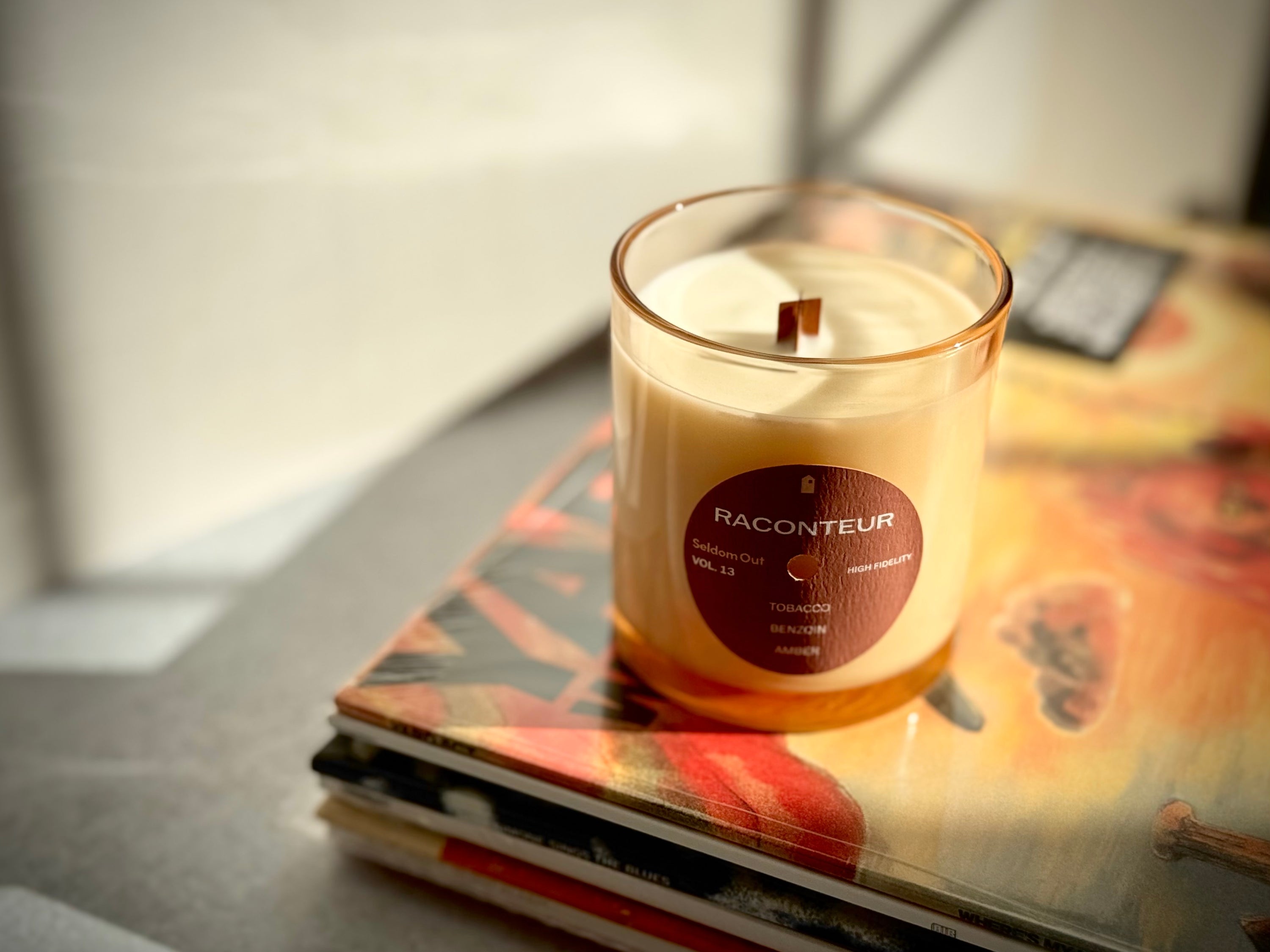 8oz Raconteur Candle in a tinted glass vessel highlighting the wooden X shaped wick at the center. The candle sits in a bright scene on top of a stack of vinyl records.