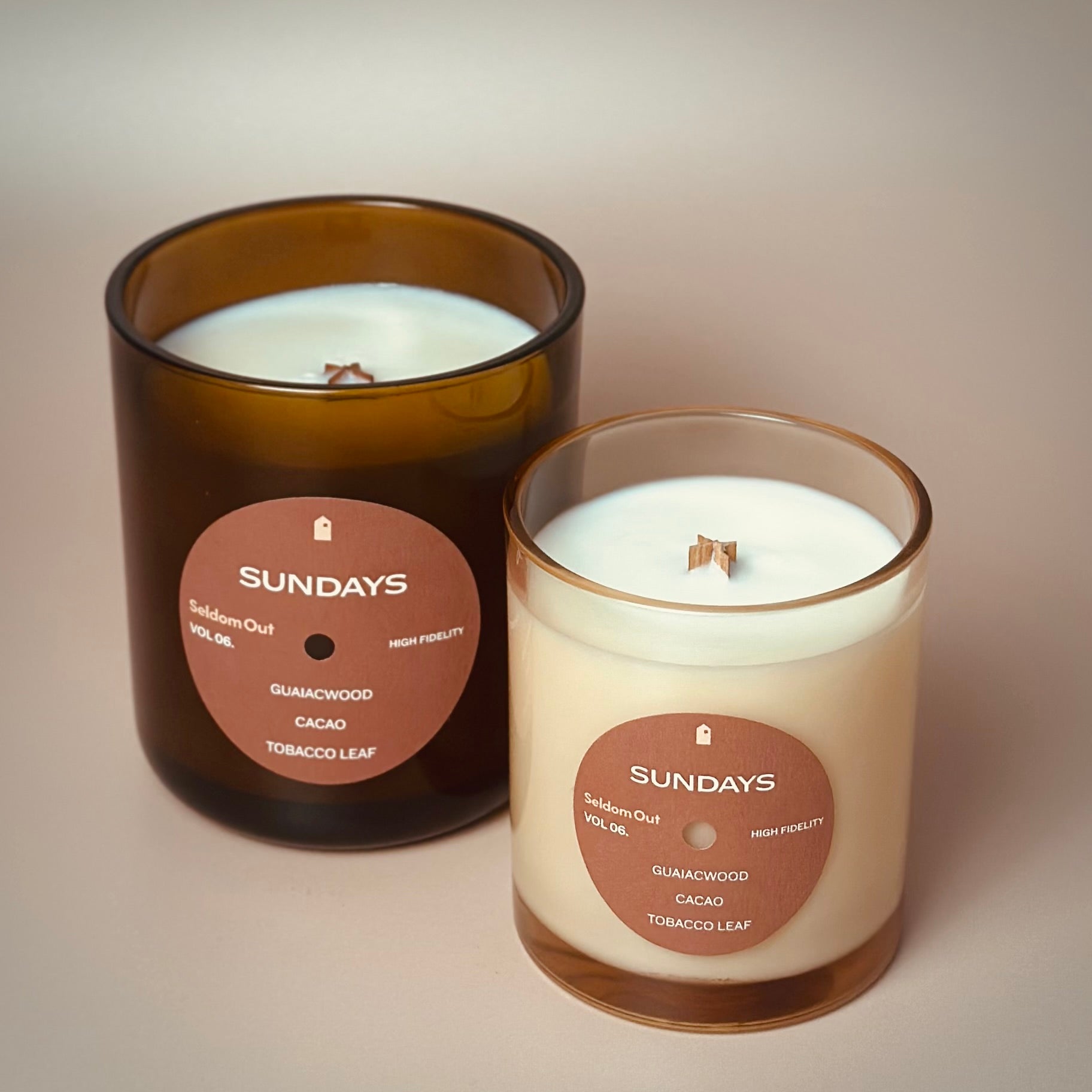 A portrait of our 8oz and 12oz Sundays scented candle standing side by side. The 8oz candle is in a lightly tinted glass vessel, the 12oz is in a heavy amber glass vessel. Both candles feature a round front label.
