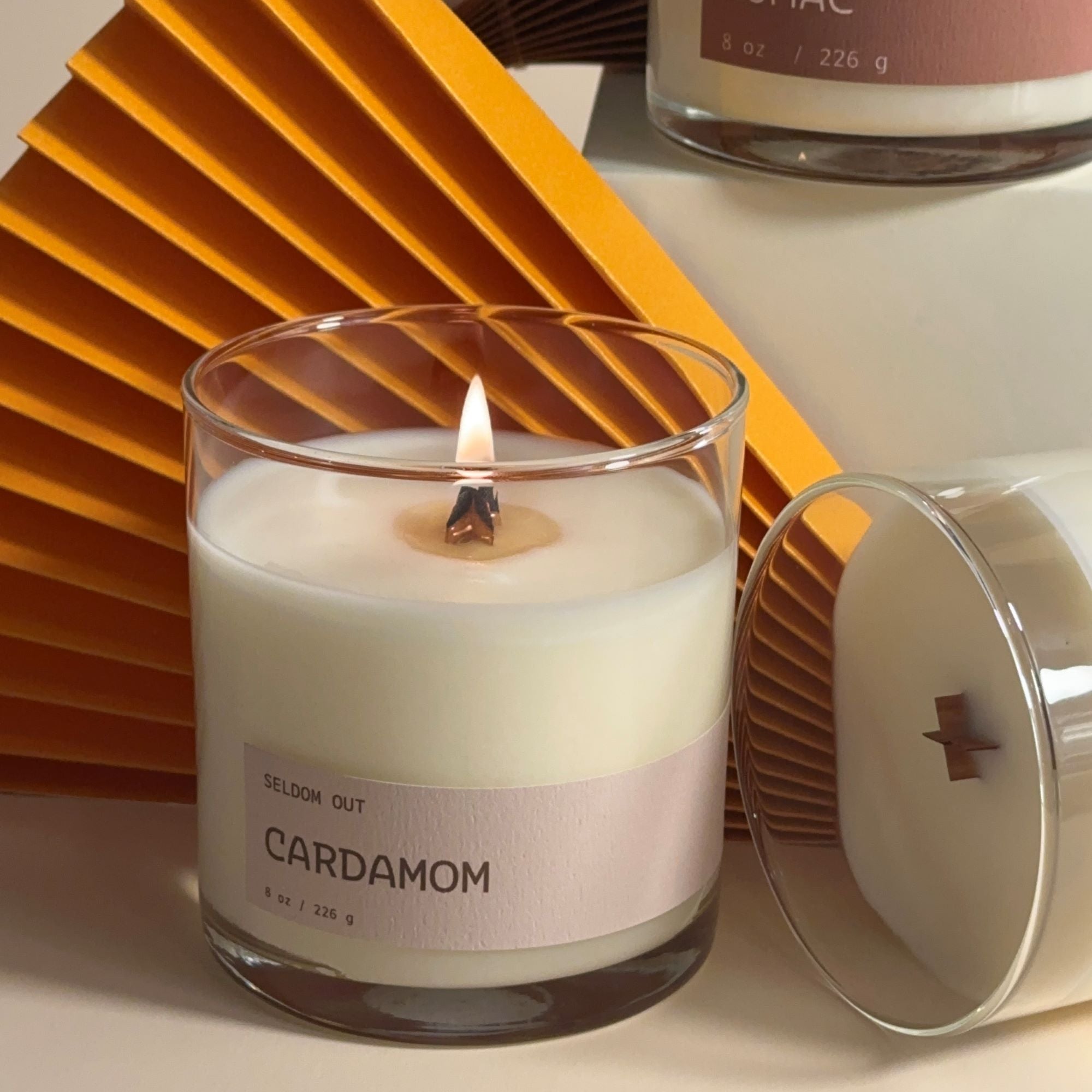 A portrait shot of our cardamom candle, with the wick alight. The vessel is a clear glass jar with a long thin label. 