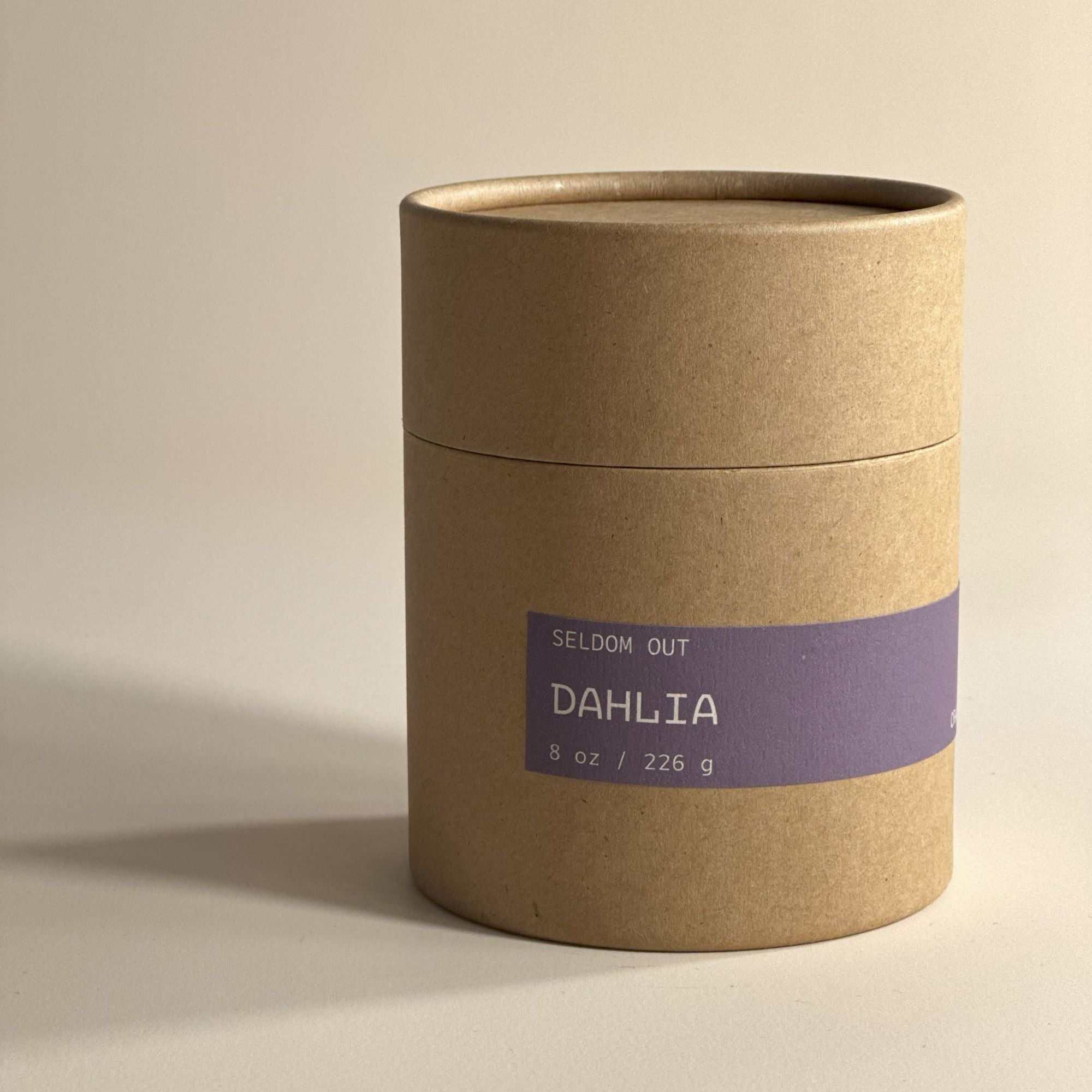 A portrait shot of the Kraft Tube packaging for our 8oz Dahlia scented candle. It features a simple long thin label. The lighting is bright but diffused with exaggerated shadows.