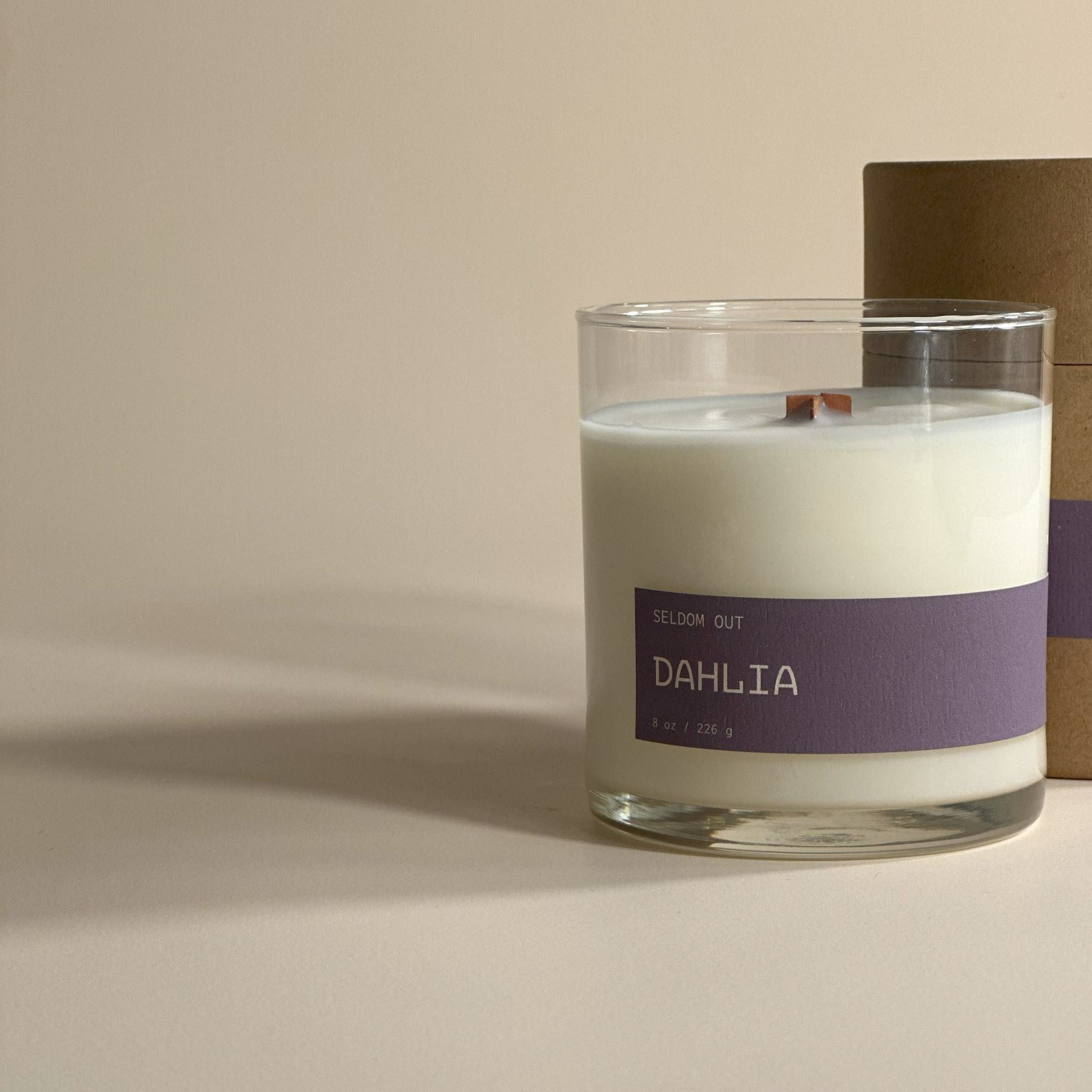 A portrait shot of our 8oz Dahlia scented candle. The vessel is a clear glass jar with a long thin label. It’s standing in front of a Kraft tube box, with a matching label. The lighting is bright but diffused with exaggerated shadows.