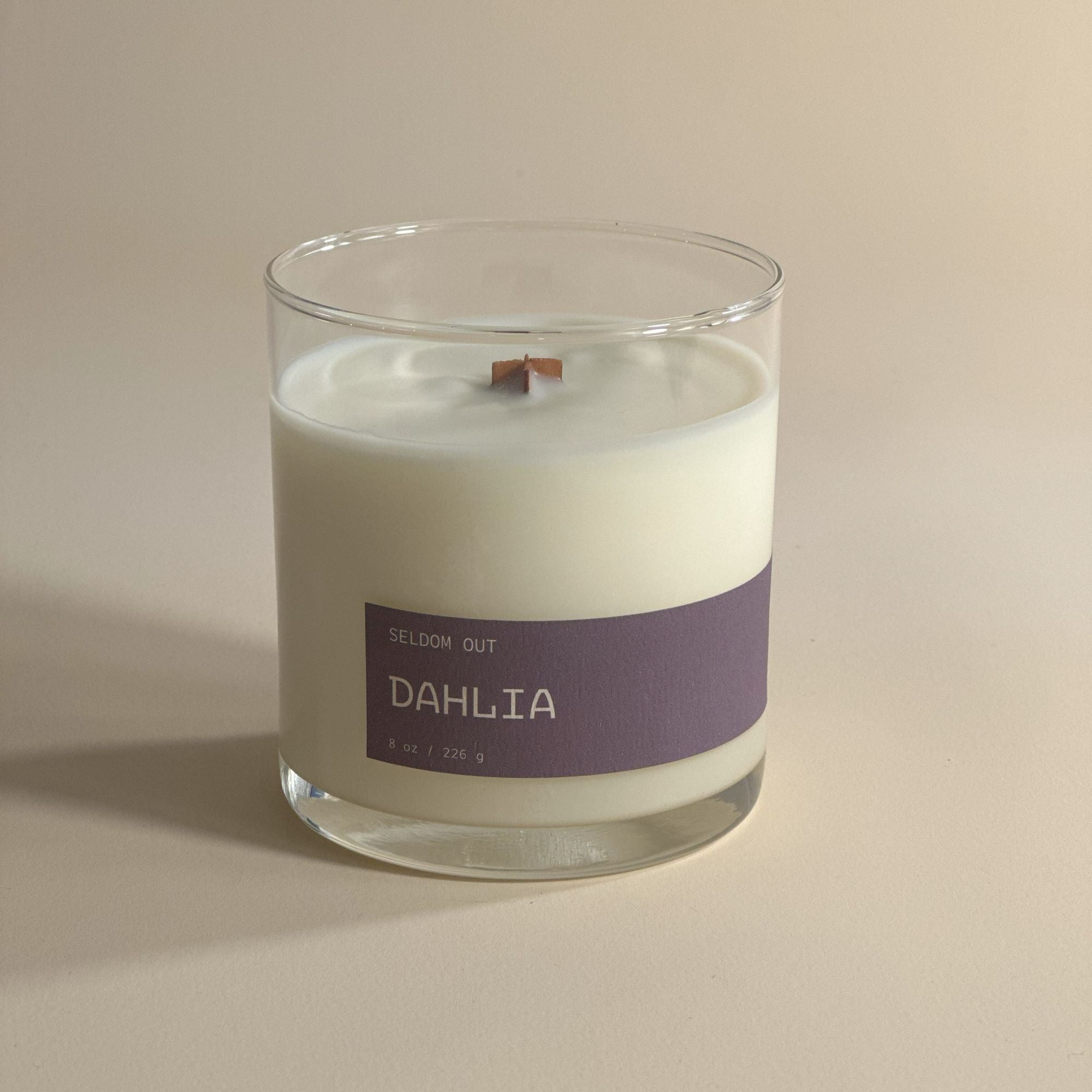 A portrait shot of our 8oz Dahlia scented candle. The vessel is a clear glass jar with a long thin label. The lighting is bright but diffused with exaggerated shadows.