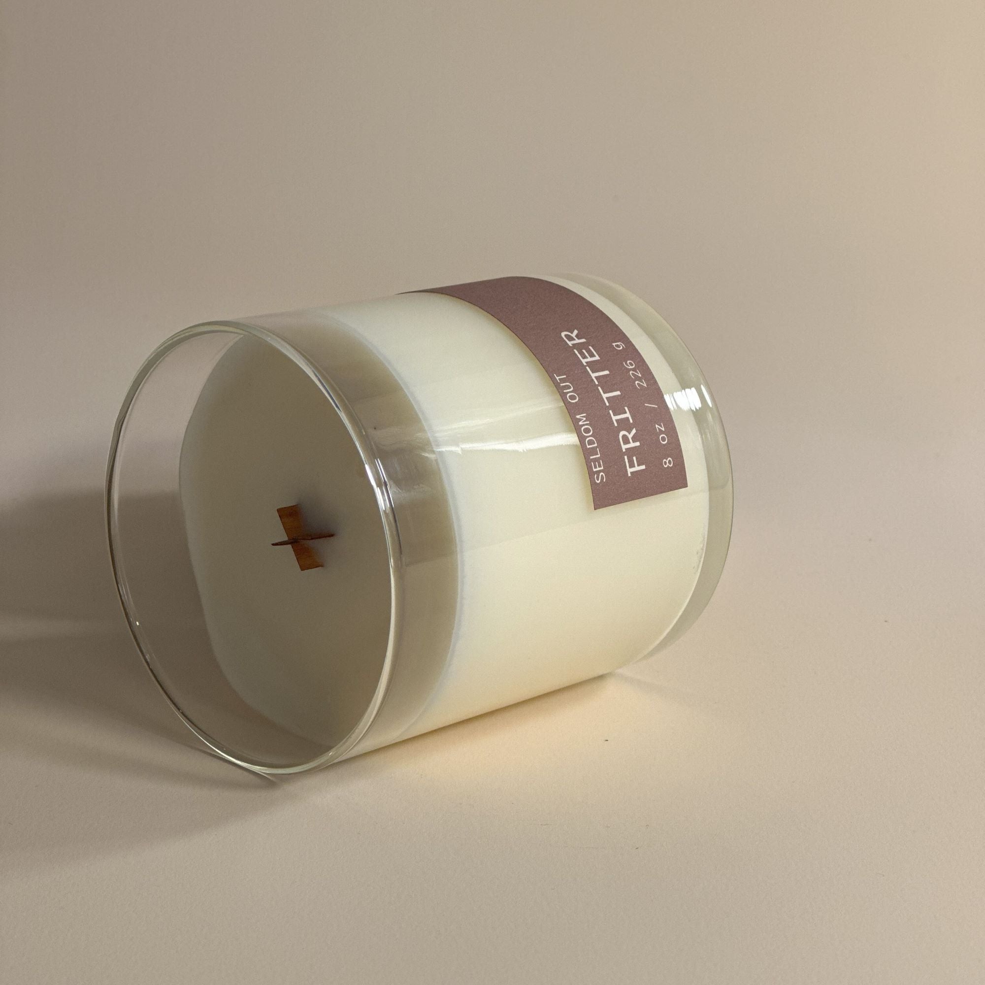 Our 8oz Fritter scented candle is shown laying on its side to highlight the wooden X-shaped wick.