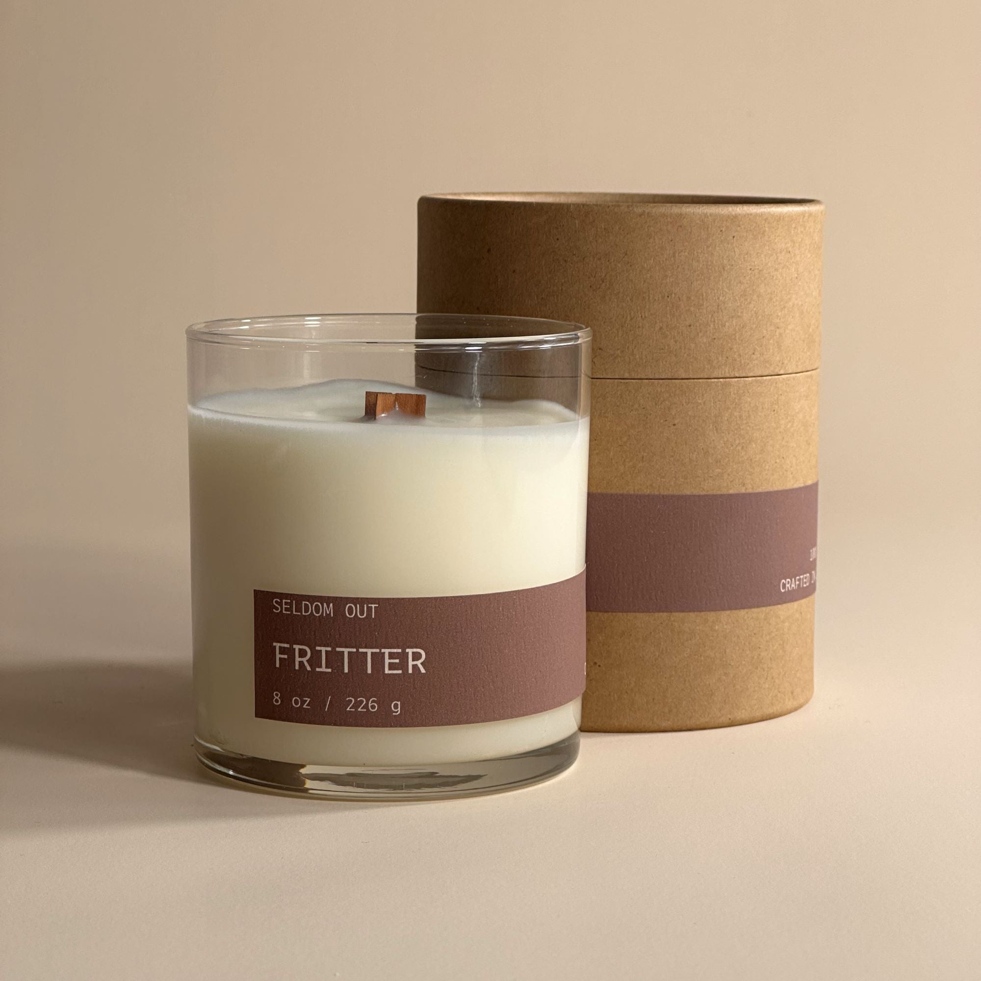 A portrait image of our 8oz Fritter Scented candle. It has a clear glass vessel, with a long thin label. It stands infront of a Kraft Tube Box with a matching label.