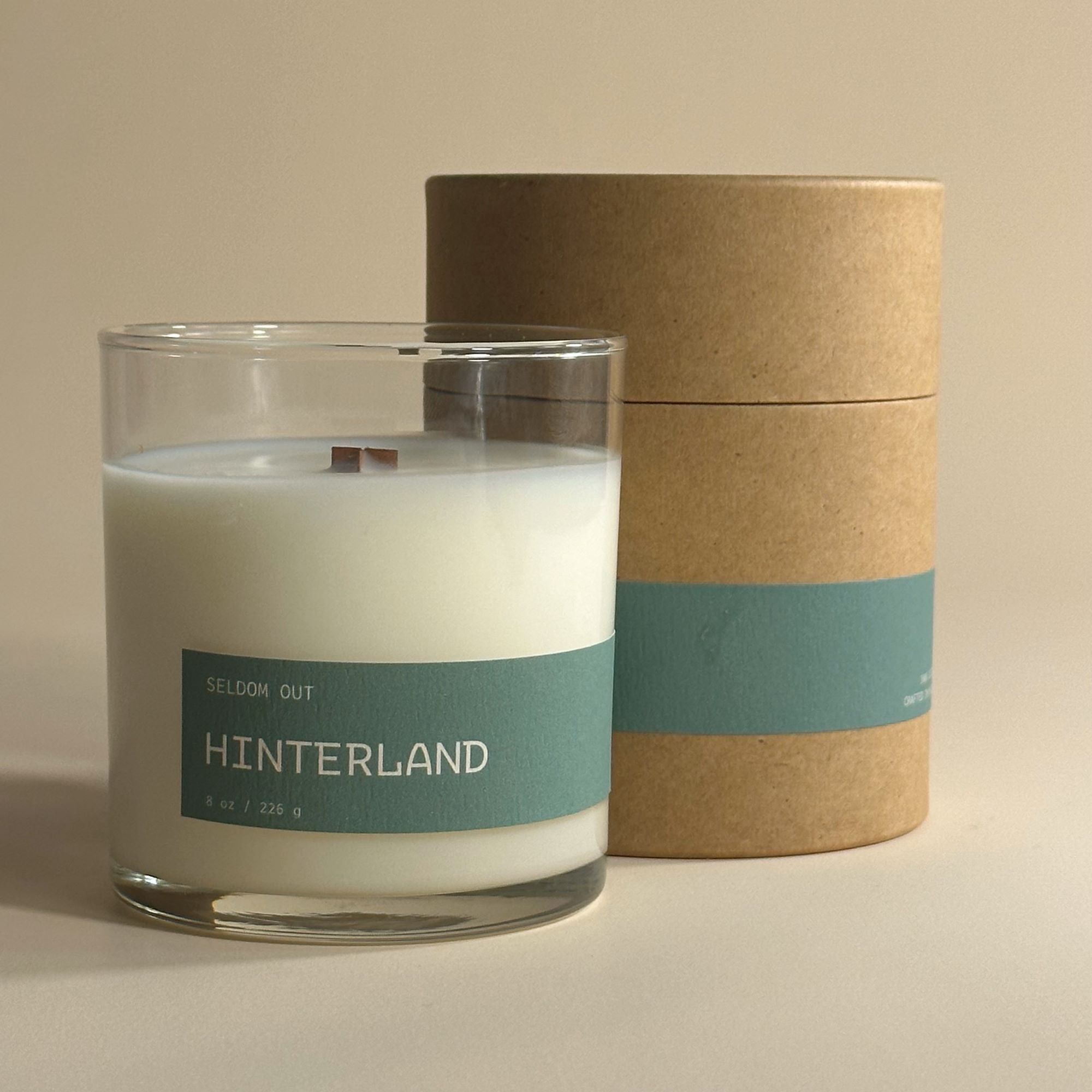 A portrait image of our 8oz Hinterland scented candle, standing in front of its Kraft tube box packaging. Both items feature matching long thin labels that wrap around the width.