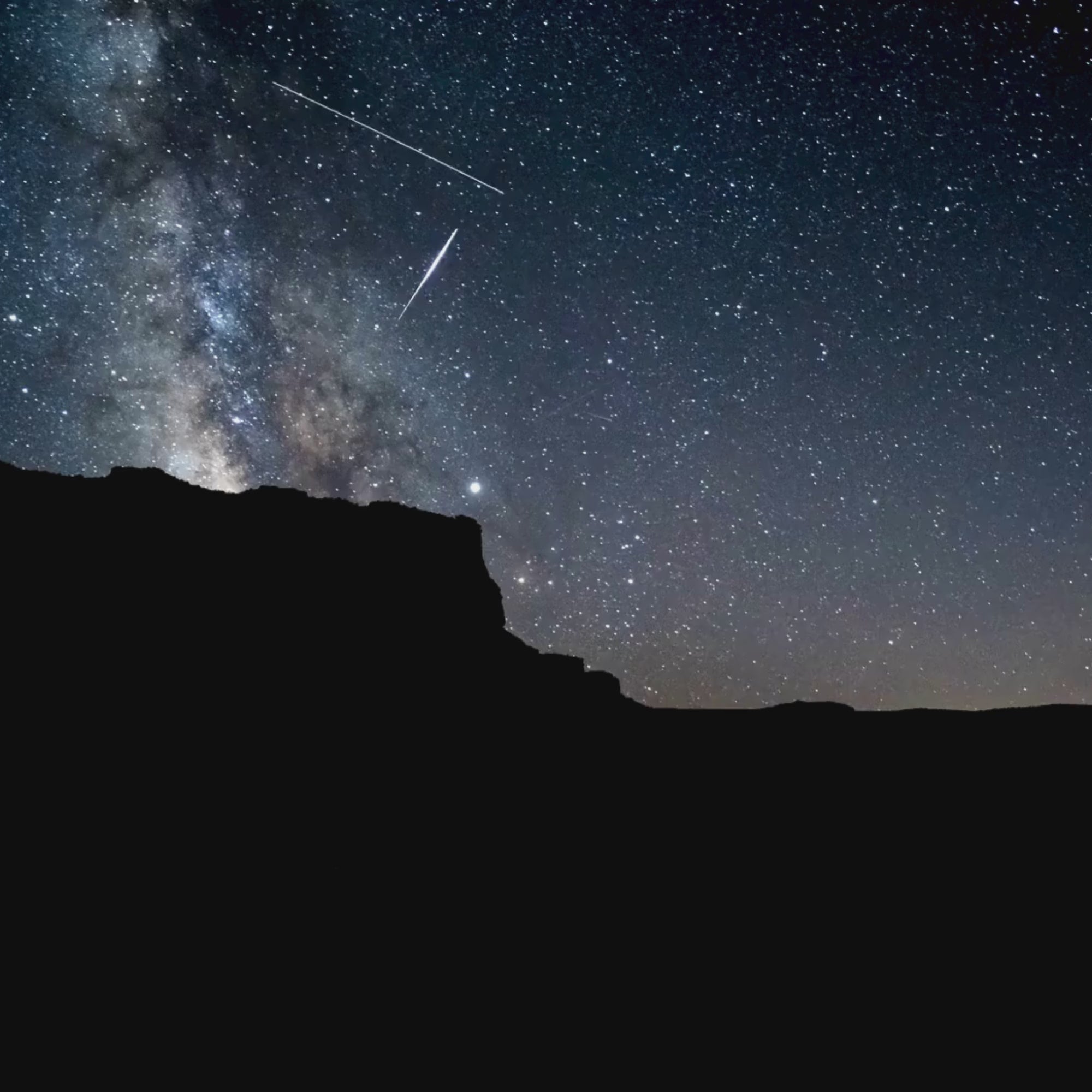 A looping video with no volume. It depicts a timelapse of a desert scene at night, with rocky terrain in the foreground and a sky  full of  thousands of twinkling stars. Some galaxies are even visible, along with shooting stars. The mood is awe inspiring.