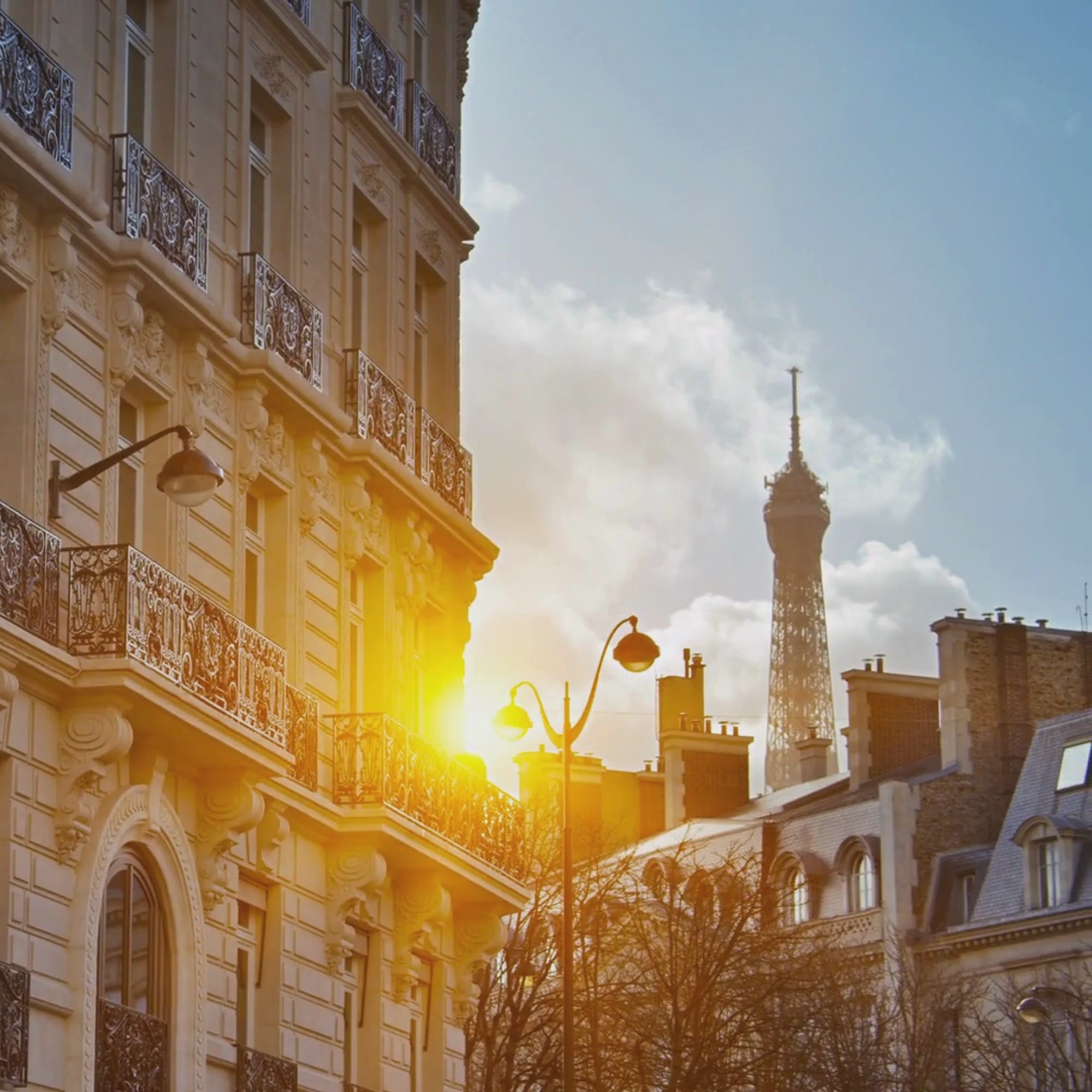 A short 5 second looping video with no volume. The video depicts a quintessential and picturesque Parisian street as the sun is low in the sky. In the foreground is a beautiful ornate building with pretty iron balconies, as the camera pans to the left, the Eiffel tower can be seen above the rooftops. The mood is relaxing.