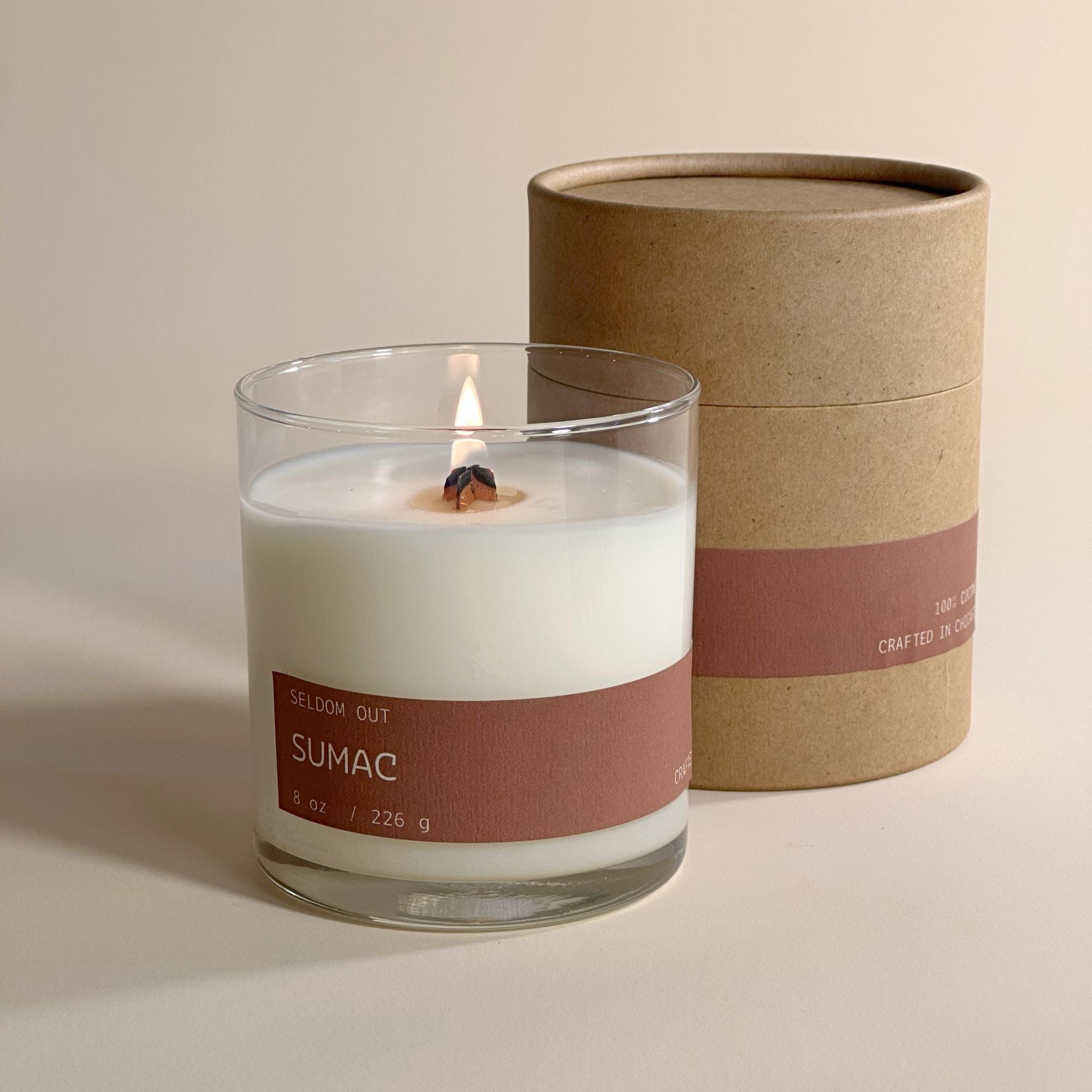 A portrait image of 8oz Sumac scented candle made in a clear glass jar, standing in-front of our packaging, a Tube box made with sturdy Kraft paperboard, both the candle & box feature matching long thin labels that wrap around the width. The wick is alight, showing a tall cylindrical flame.