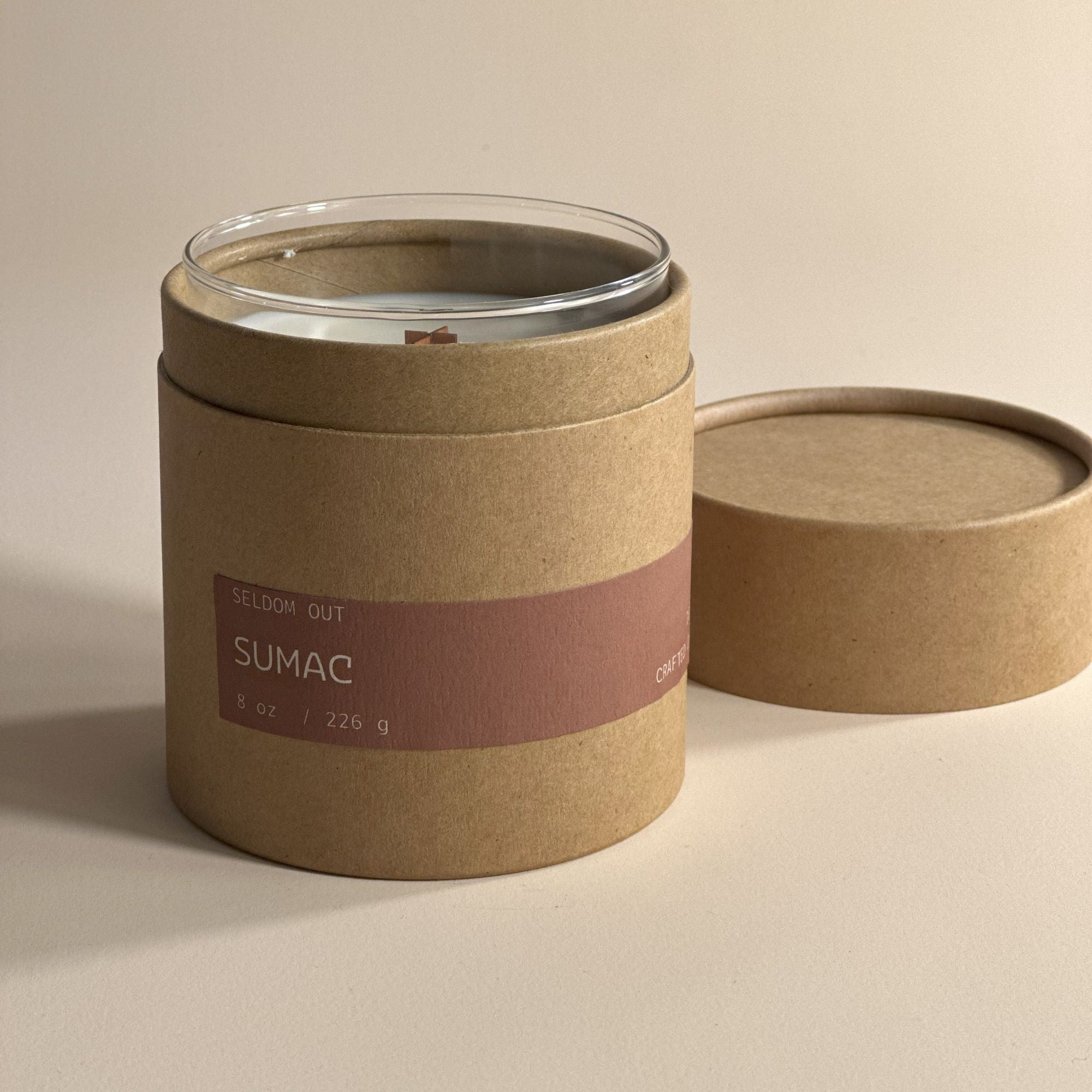 A portrait image of the tube box for our 8oz Santal scented candle. Made with sturdy Kraft paperboard it features a long thin label. The lid has been removed and sits to one side, a hint of the clear glass vessel is visible.