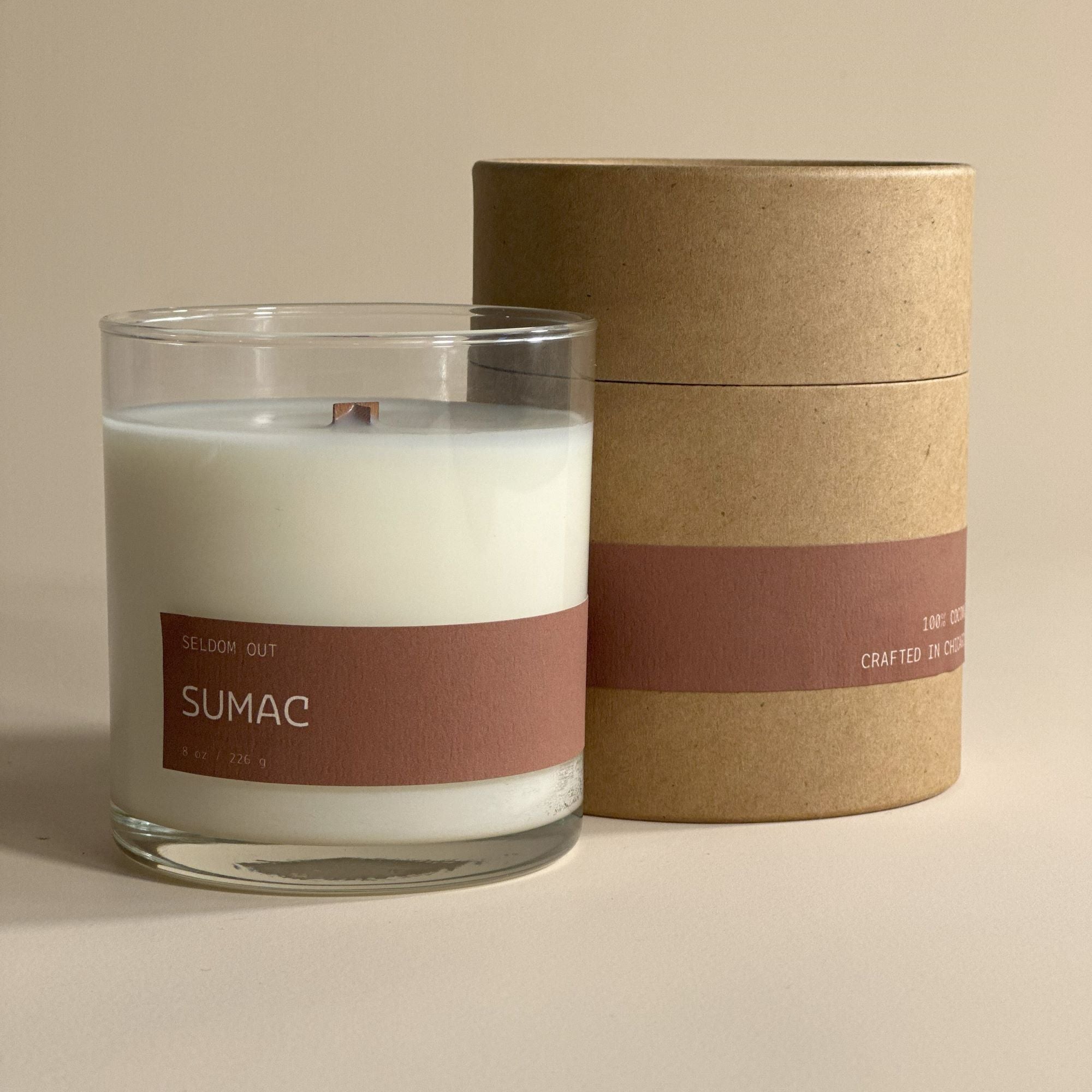 A portrait image of our 8oz Sumac scented candle made in a clear glass jar. Behind it stands its box, a tube made with sturdy Kraft paperboard. Both the candle and the box feature matching long thin labels that wrap around the width.