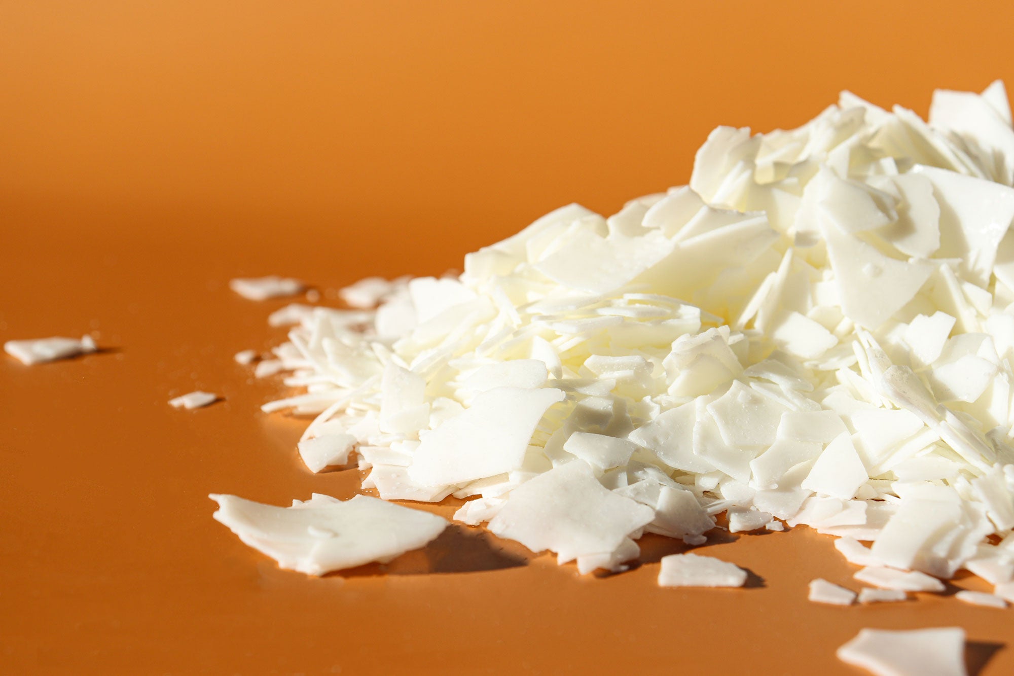 A close up image of creamy white wax flakes arranged in a pile against a vibrant orange background.